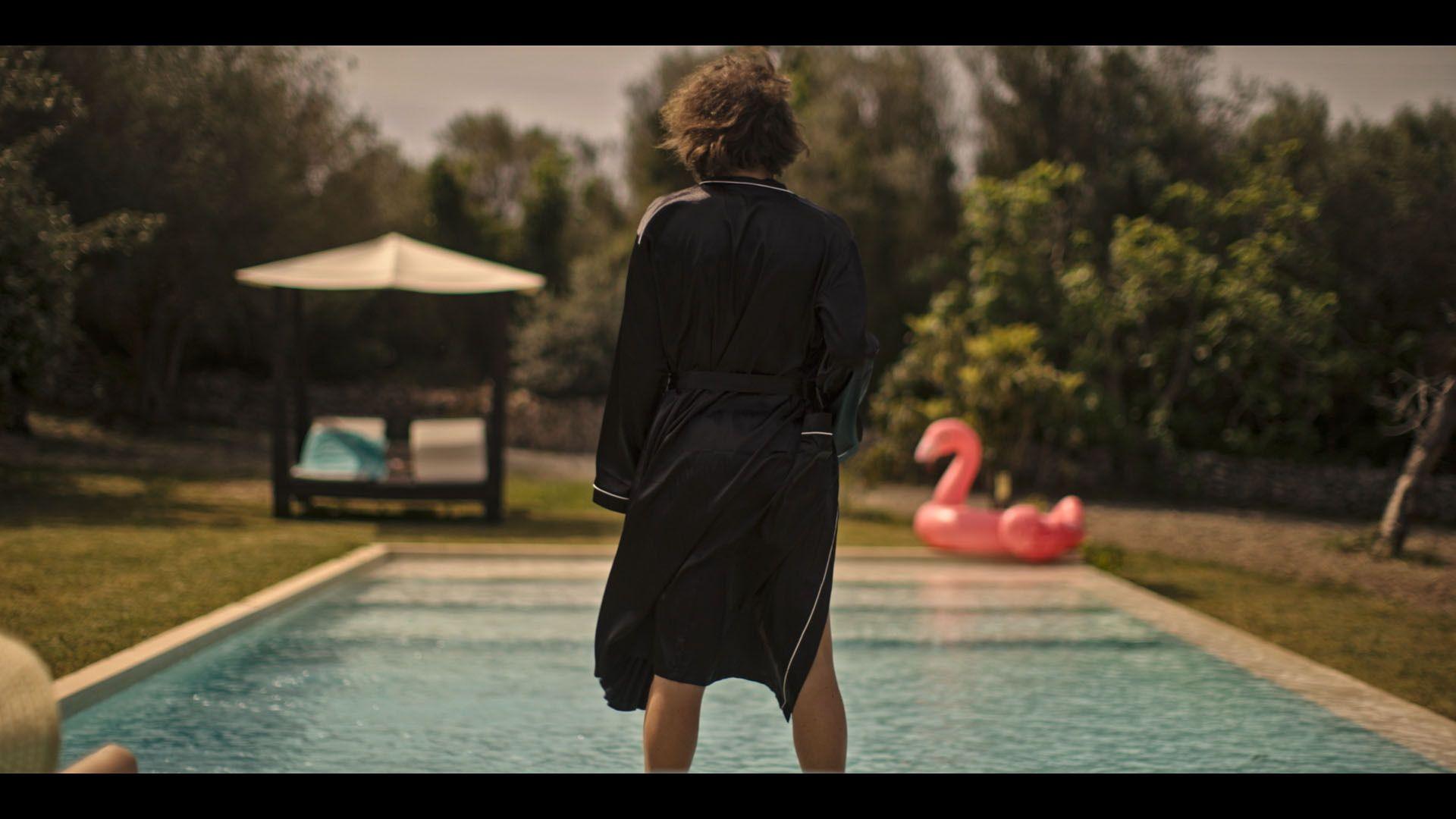 A reconstruction from the documentary shows Howard Marks (played by Diego Marando), shot from behind, wearing a black dressing gown and looking at a swimming pool with a blow up flamingo. 