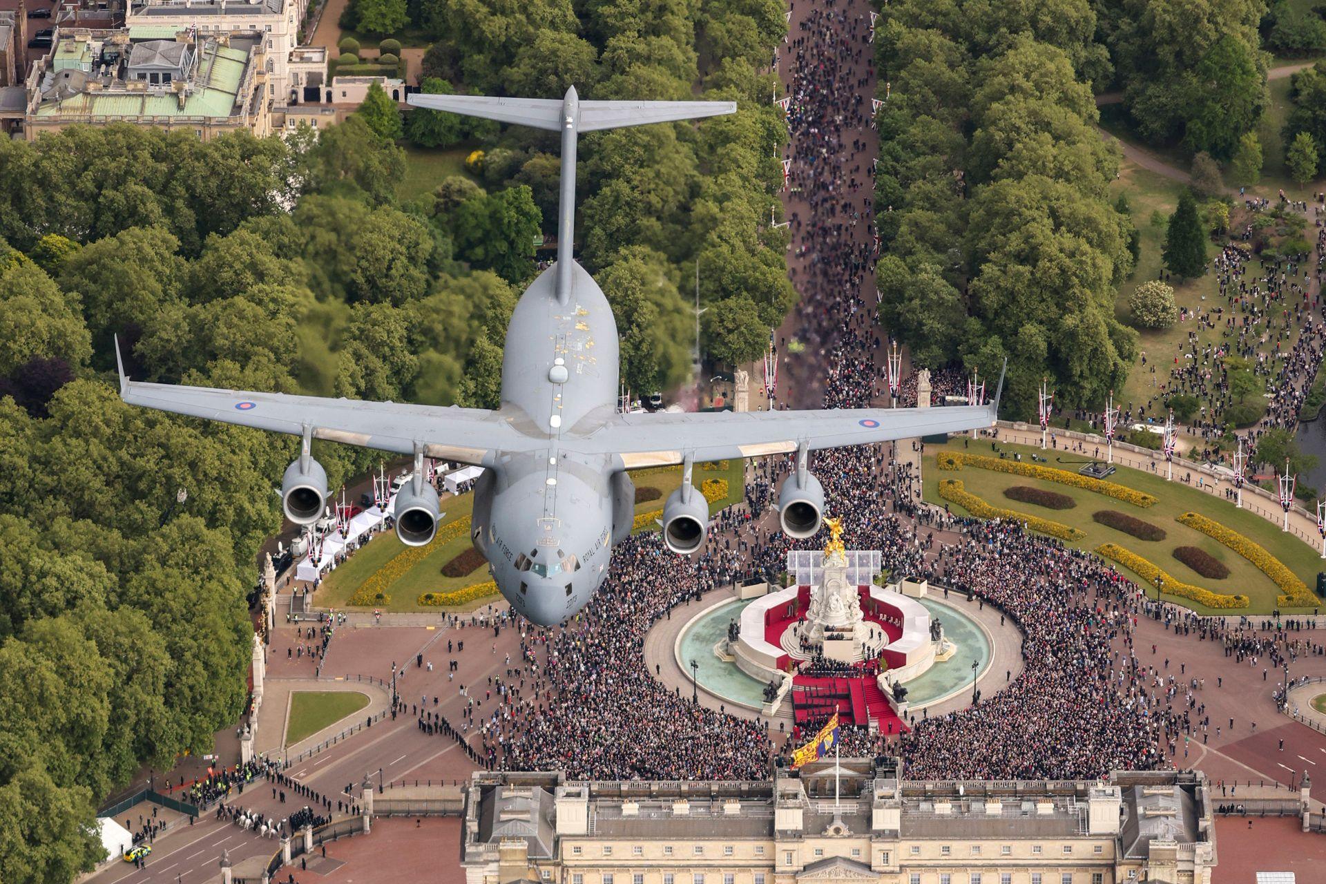 A grey C17 Globemaster bomber plane is pictured flying through the air, having been captured by a photographer positioned on another aircraft above it which is out-of-frame. Below the aircraft a huge crowd is seen gathered around the statue and fountain in front of Buckingham Palace with green foliage around them.