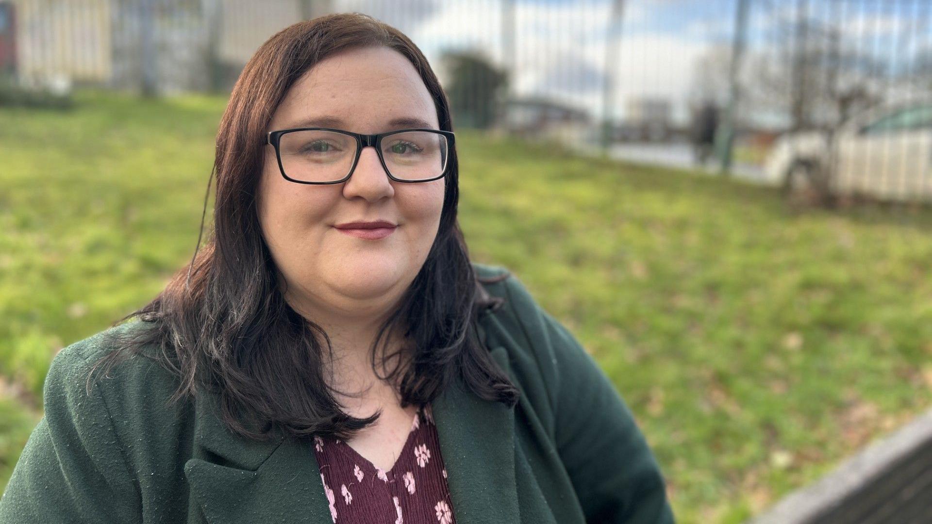 Labour councillor Hazel Dawkins is wearing a green coat, a red patterned top and glasses. She is smiling at the camera. The background is blurred but she is near a grassy verge on a road with a metal fence behind. 