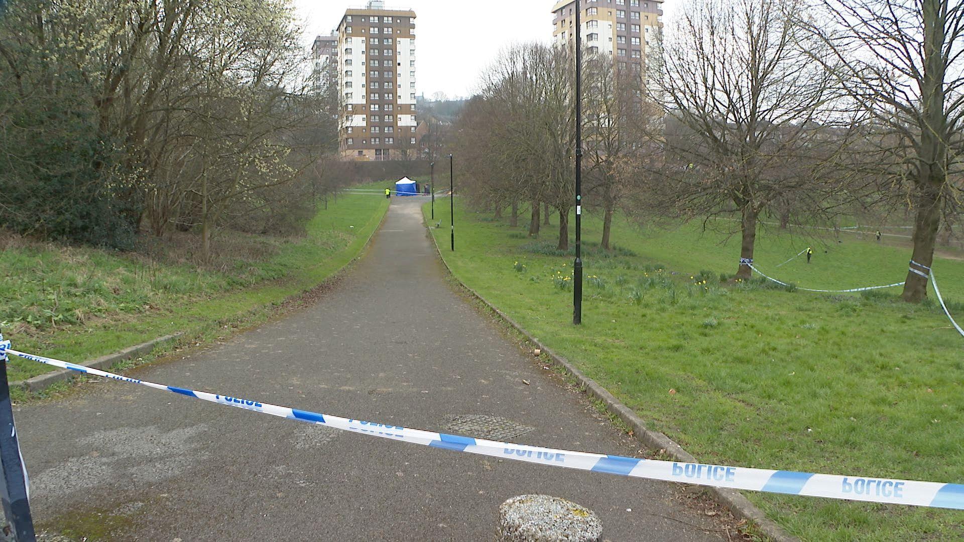 A long grey path flanked by two patches of grass. There is a blue tent in the background, near some blocks of flats. There is blue and white police tape in the foreground