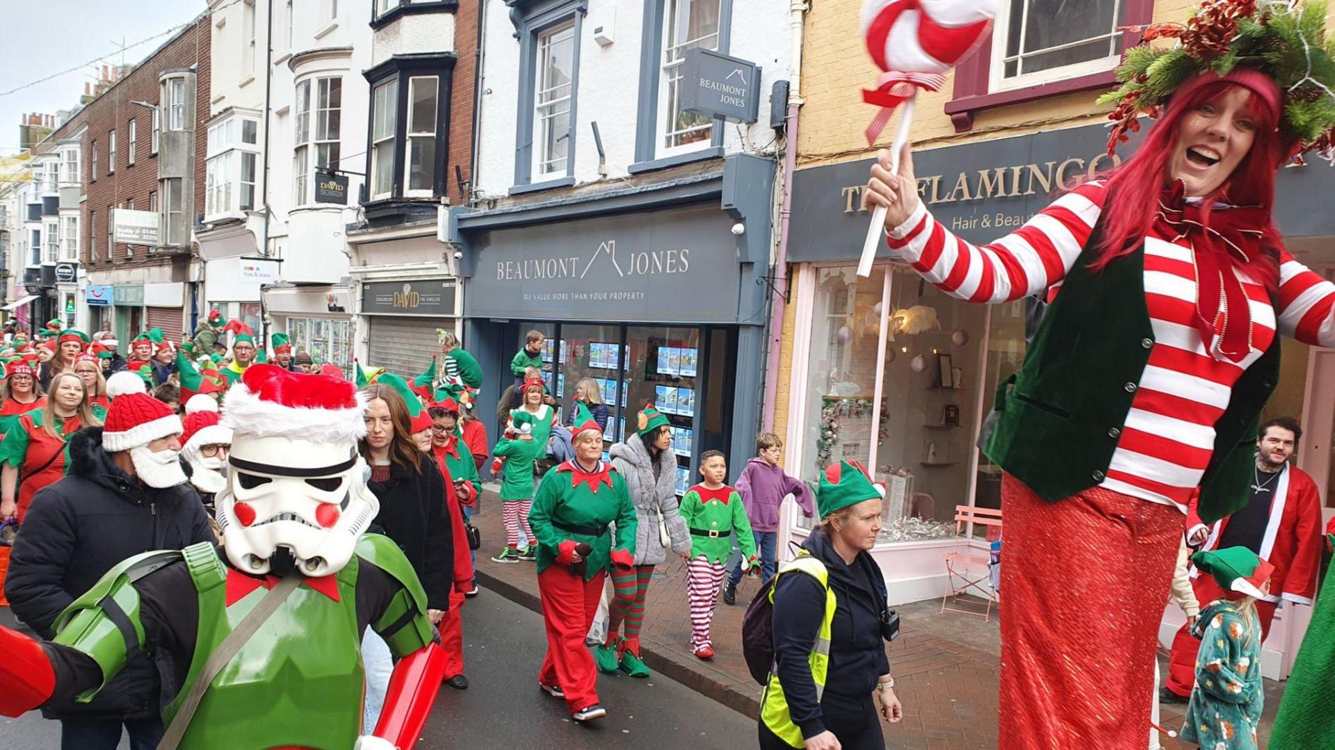 Hundreds of people dressed as elves walking along a shopping street in Weymouth. In the front of the picture is a Star Wars stormtrooper dressed as an elf with red rosy cheeks stuck on the white stormtrooper helmet.