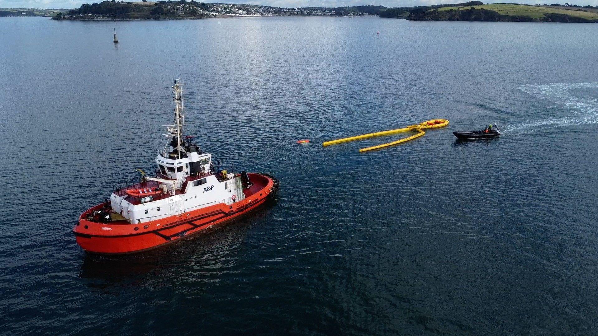 A drone shot of the response to the mock oil spillage. There is a large and tall orange and white boat with a smaller black boat behind it. Directly next to the black boat and behind the larger orange boat is a yellow inflatable item.