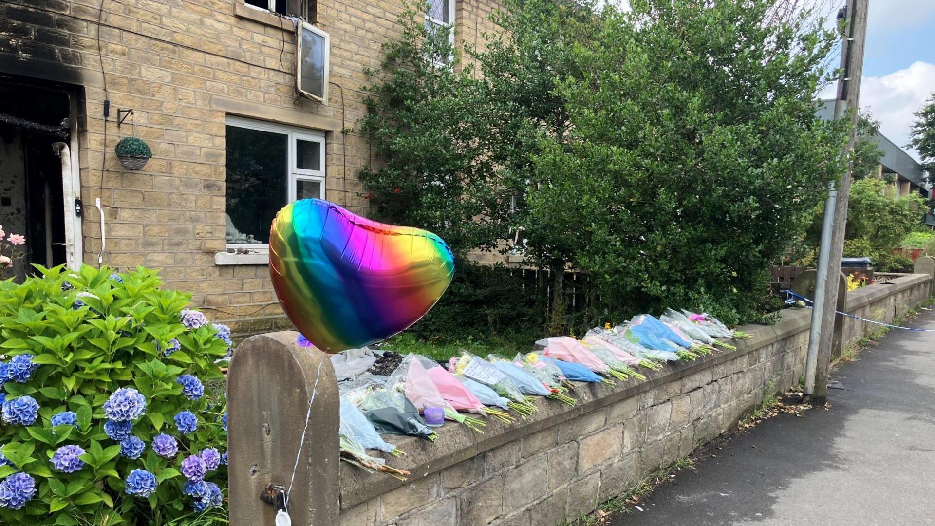 A rainbow coloured balloon and bunches of flowers left on a wall outside a burned-out house