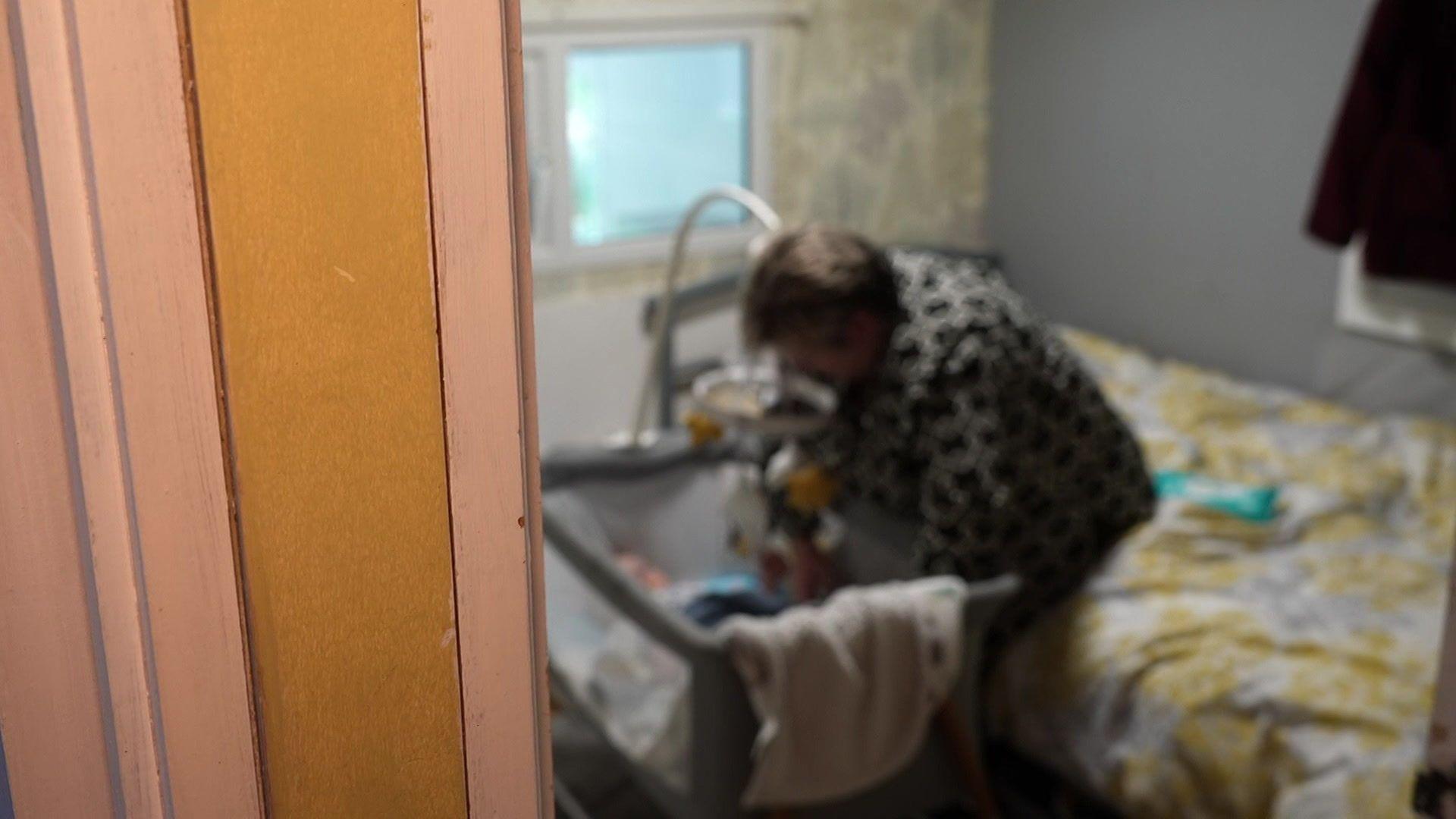 A woman is in a bedroom, sitting on a bed and placing a young baby into a cot