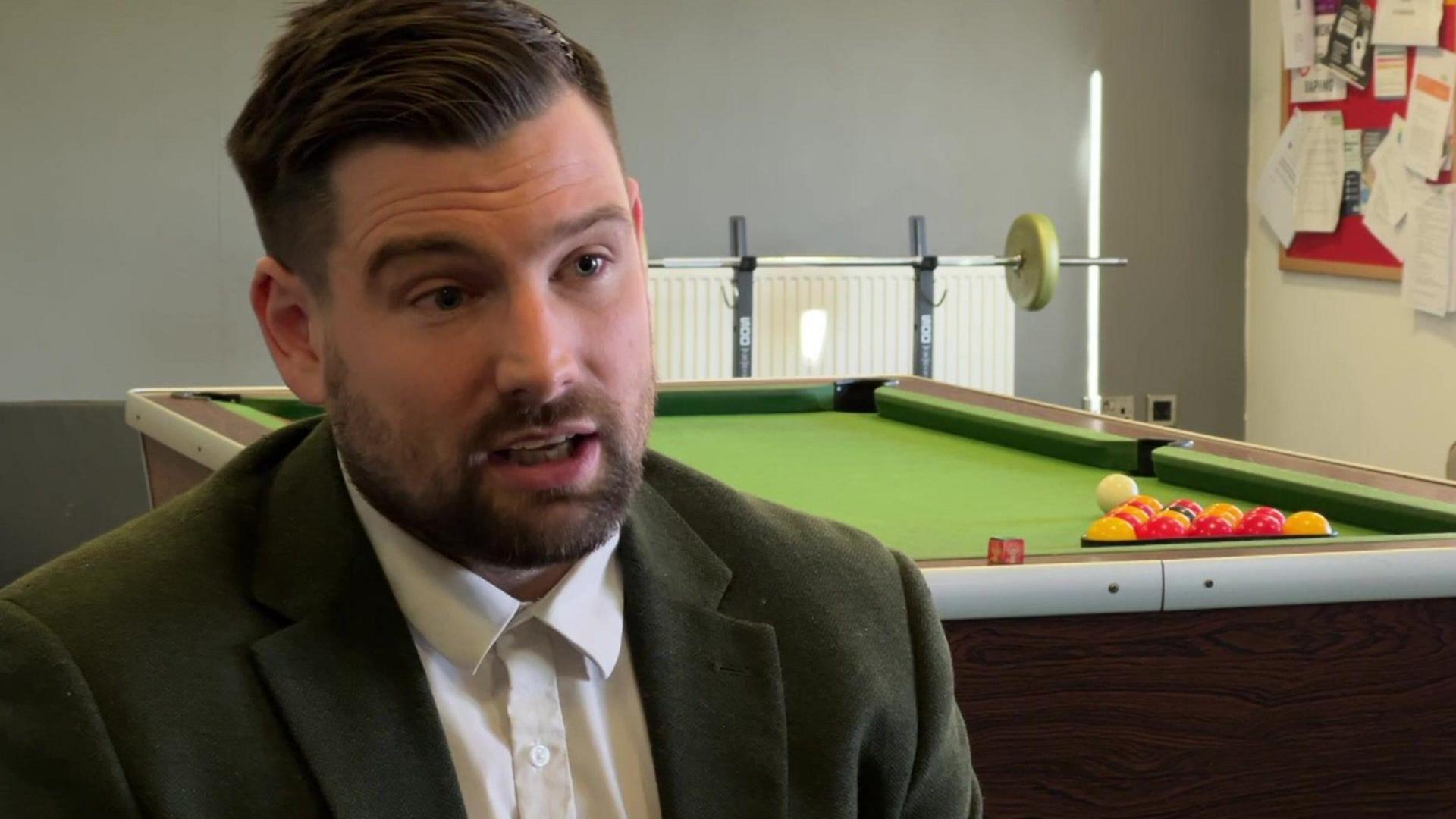 A man with short dark hair and a trim beard talking to a camera. He is wearing a dark green jacket and a white shirt and is sitting on a chair in a room with a pool table and a weights bench behind him