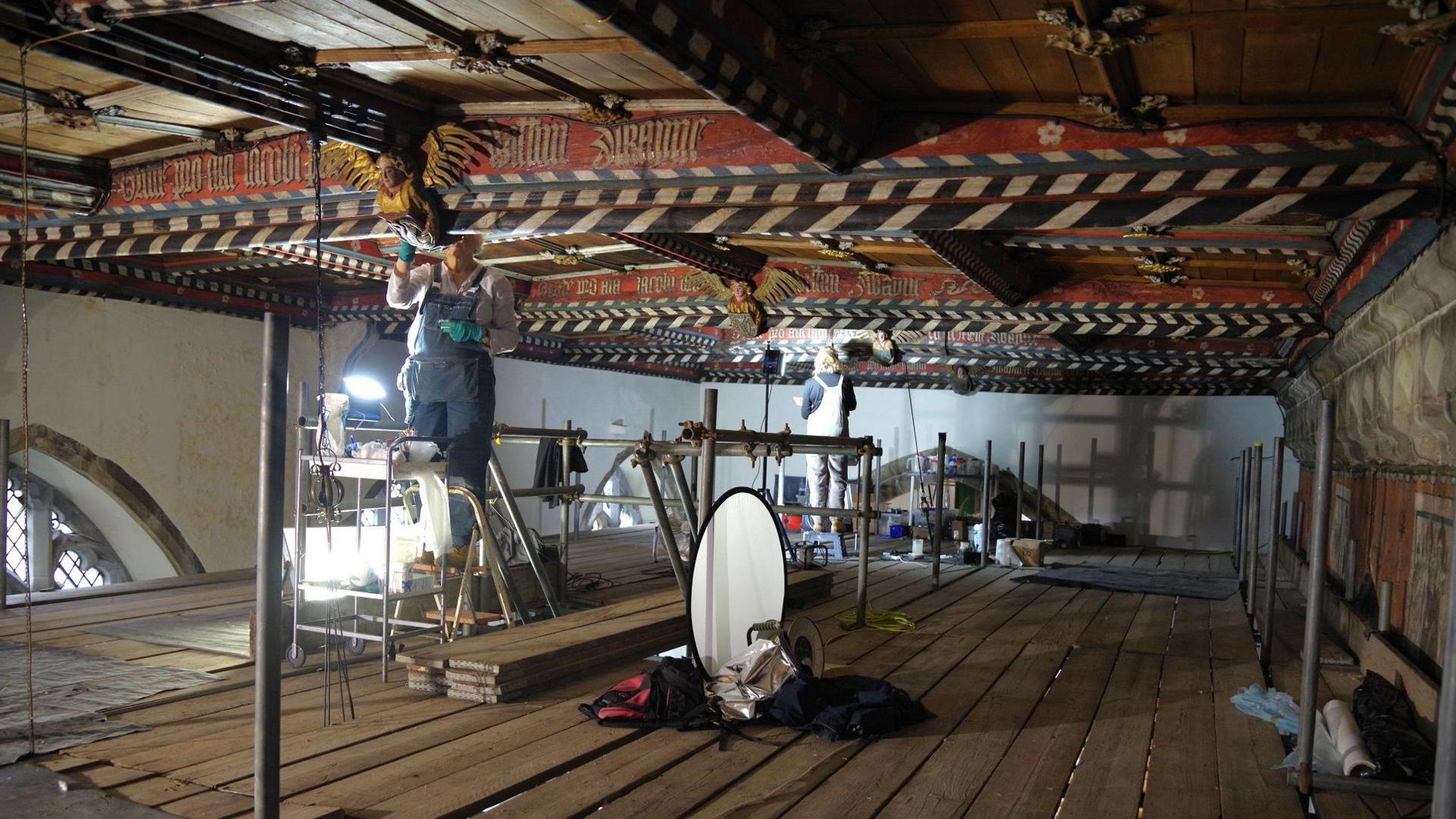 A view of what could be an attic with an elaborate coloured ceiling, but is lots of scaffolding boards with equipment such as lighting and step-stools in the rafters of a church. Two people at work carefully looking at beams.