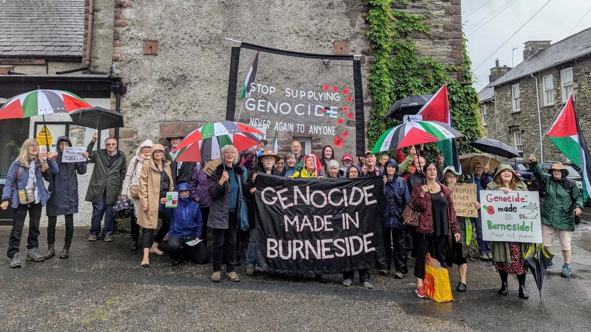 Group of people standing outside holding Palestine flag coloured umbrellas and signs which say: "Stop supplying genocide - never again to anyone" and "Genocide made in Burneside"