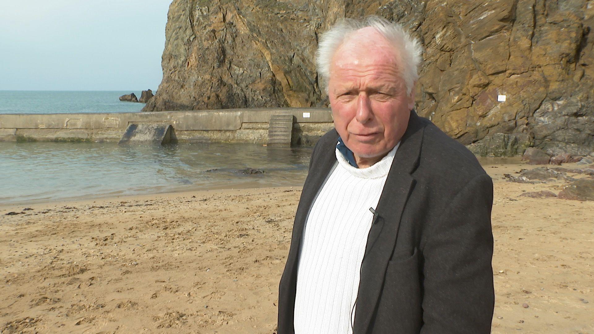 The Comic Strip actor and director Peter Richardson at Hope Cove standing in front of the breakwater on a sunny day. He has white hair and is wearing a cream jumper and a dark grey jacket.