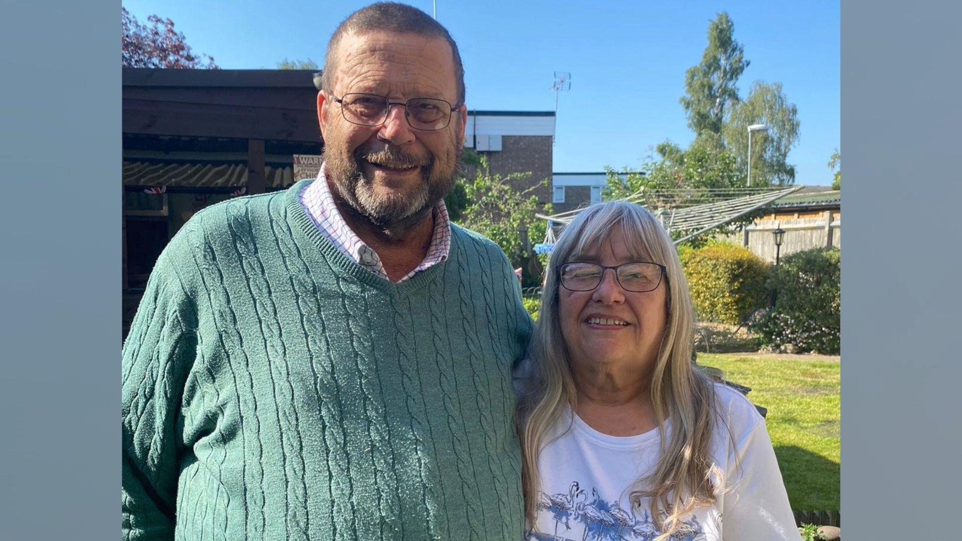 Kevin and Sue Starling. Kevin, on the left, appears in a checked shirt and green jumper over the top. His wife also wears glasses, and has a white t-shirt on. They are standing in a garden