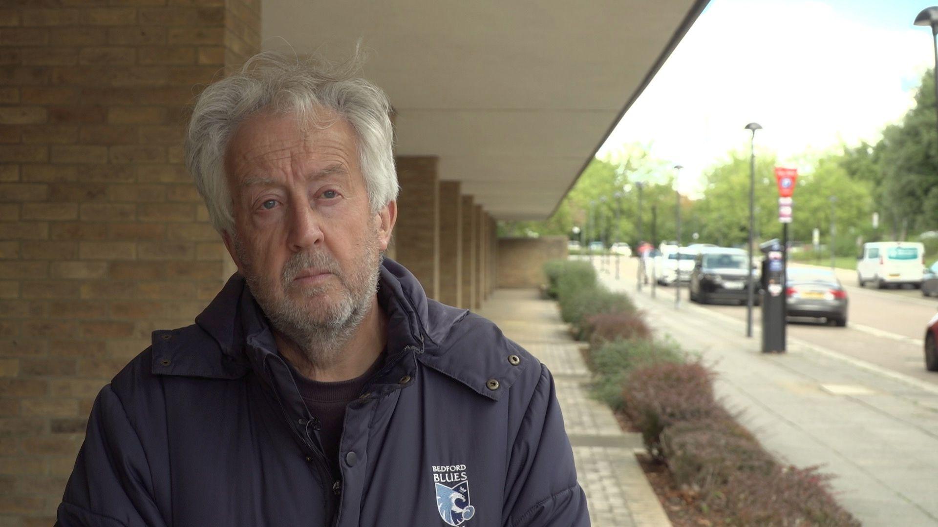 A medium close-up of David Hopkins, who is looking to the right of the camera. He has grey hair and a short greying beard. He is wearing a blue "Bedford Blues" coat, over a dark top . Mr Hopkins is standing outside a building. In the background are several brick pillars and a row of low level hedges. To the right of the image are several parked cars.