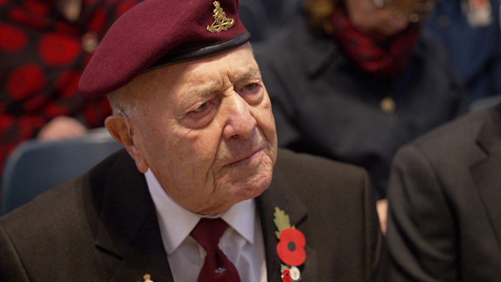Geoff Mitchell wears a maroon beret and a suit and maroon tie. He has a poppy on his lapel. He is leaning forward with a pensive look on his face.