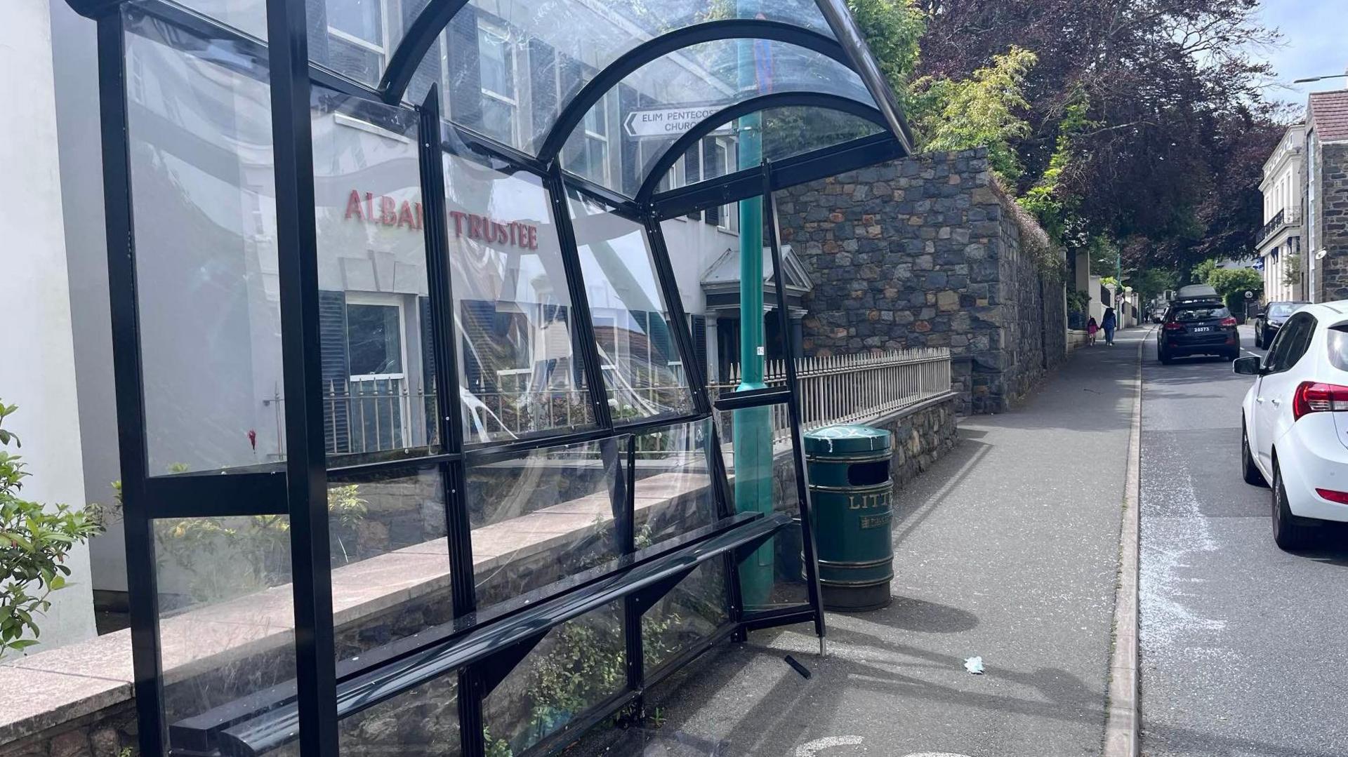 A glass bus stop. One end is at an angle and shattered glass sits over the road.