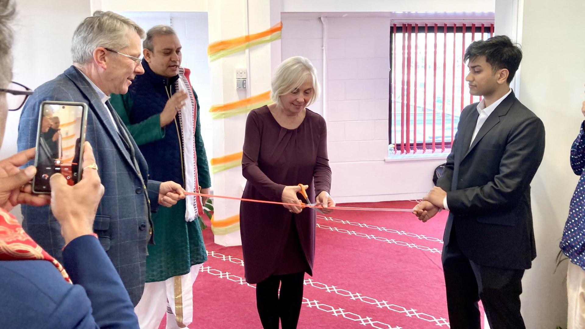 A woman cutting a red ribbon to symbolise the official opening of the temple, while two men on either side hold it taut. The room is decorated with orange and yellow ribbons wrapped around a column, and there is someone taking a picture of the ribbon cutting on the left. The carpet is red and the room is bright.