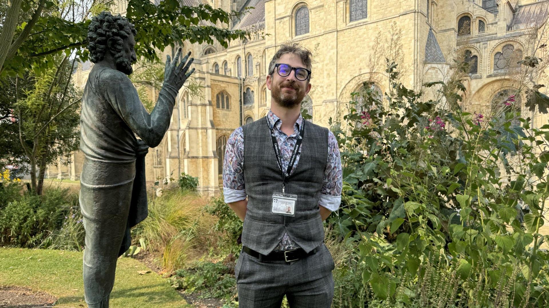 Simon Yule, the cathedral's visitor experience manager, dressed in a waistcoat and matching trousers, standing beside a statue in the precincts of Canterbury Cathedral.