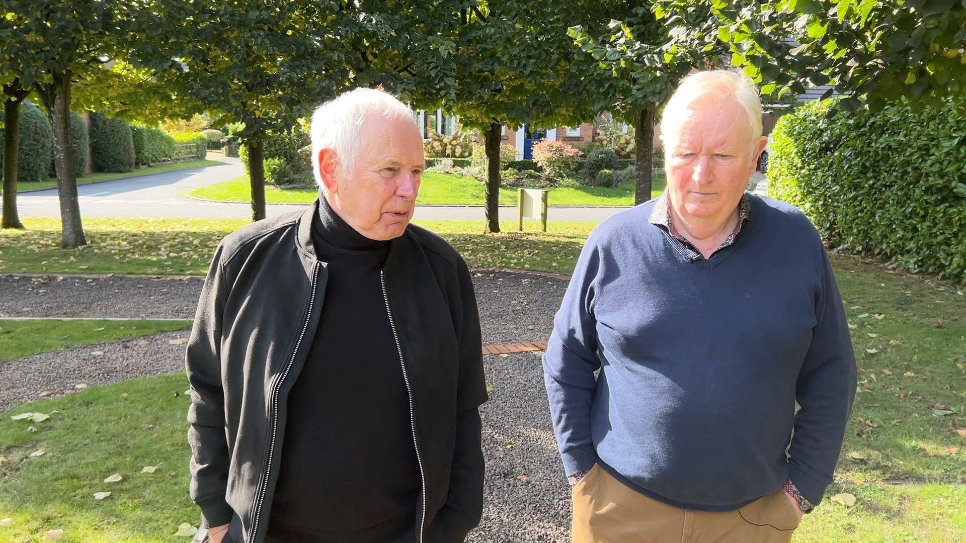 Nick Owen and Ian Phillipson walking along a street together. Nick is wearing a black jacket and black turtle-neck jumper. Ian is wearing a blue jumper over a colourful shirt.