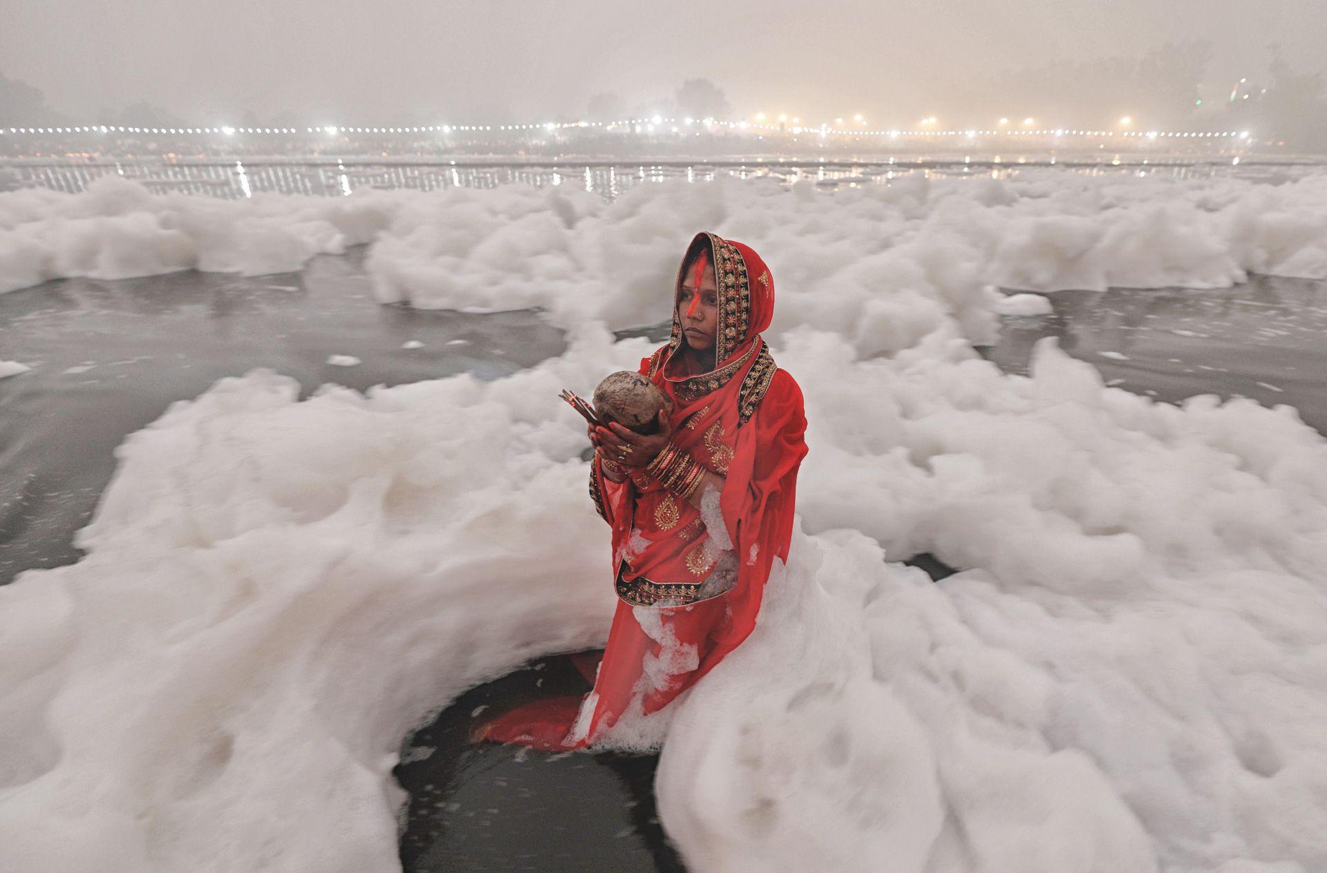 A devotee stands in the Yamuna River, New Delhi, offering prayers to the sun, as the water - thick with foam - surrounds her. She is wearing a red sari