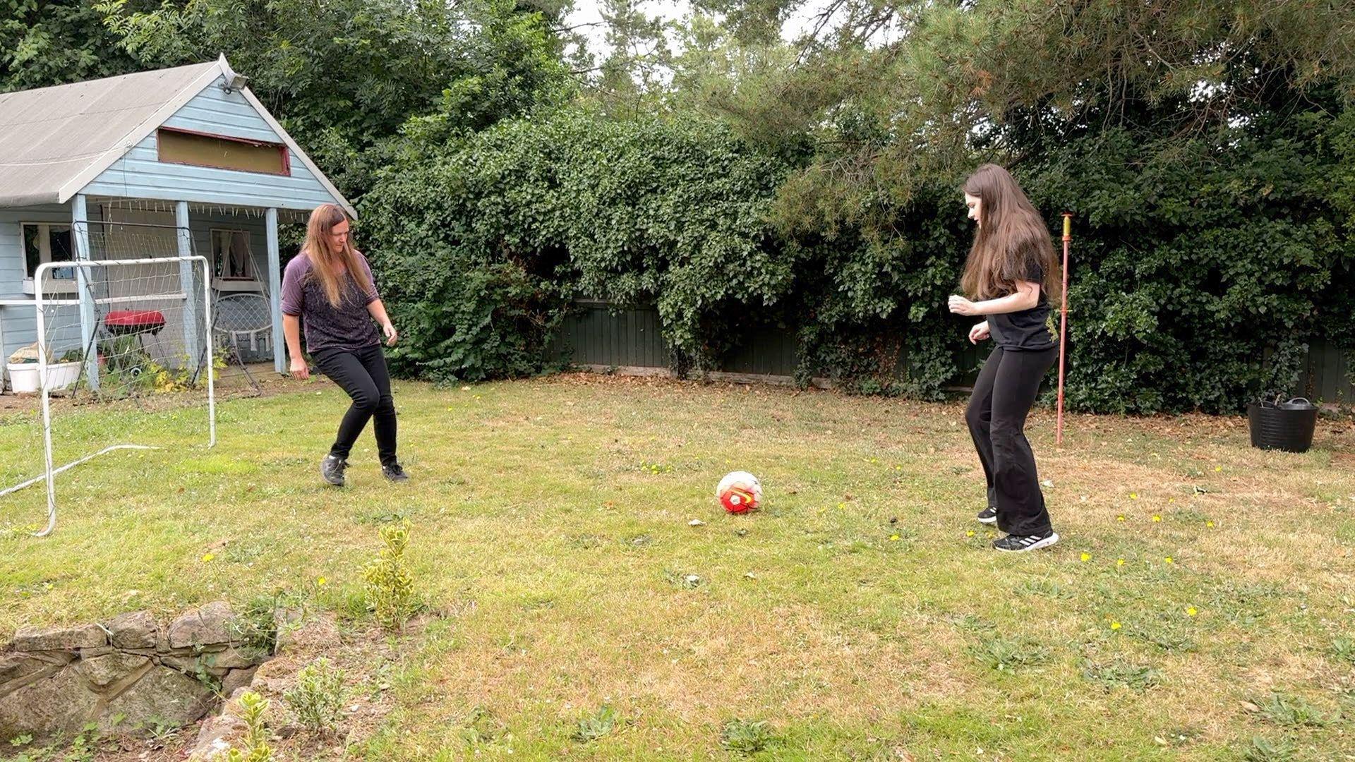 Two men women kick a ball around a garden. A shed and goalposts can be seen in the background, alongside trees.