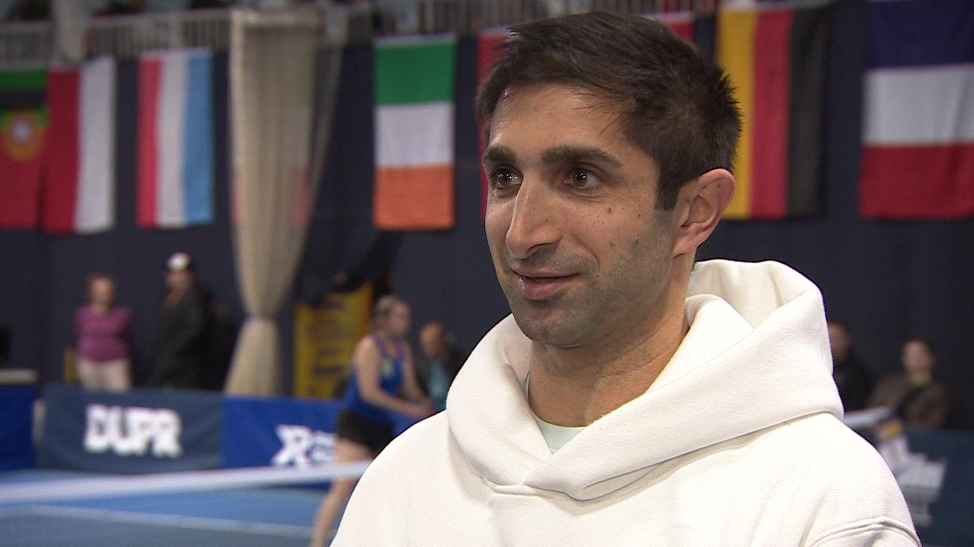 James Chaudry giving an interview. He is a wearing a white hoody. He is looking away from the camera. The flags of various European countries are seen hanging in a wall behind him. One player could also be seen on the court.