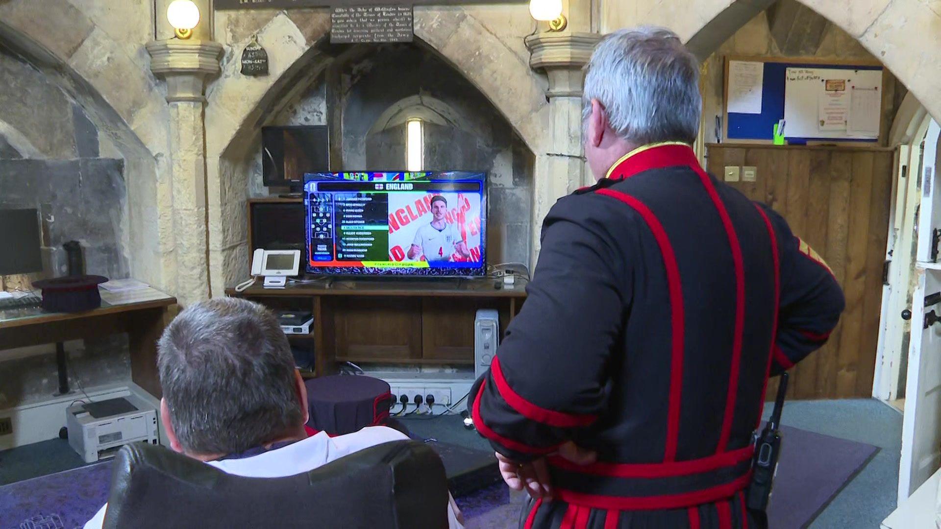 Two men watch a television showing an England football screen inside a stone-walled room with arched ceilings and wooden furniture.