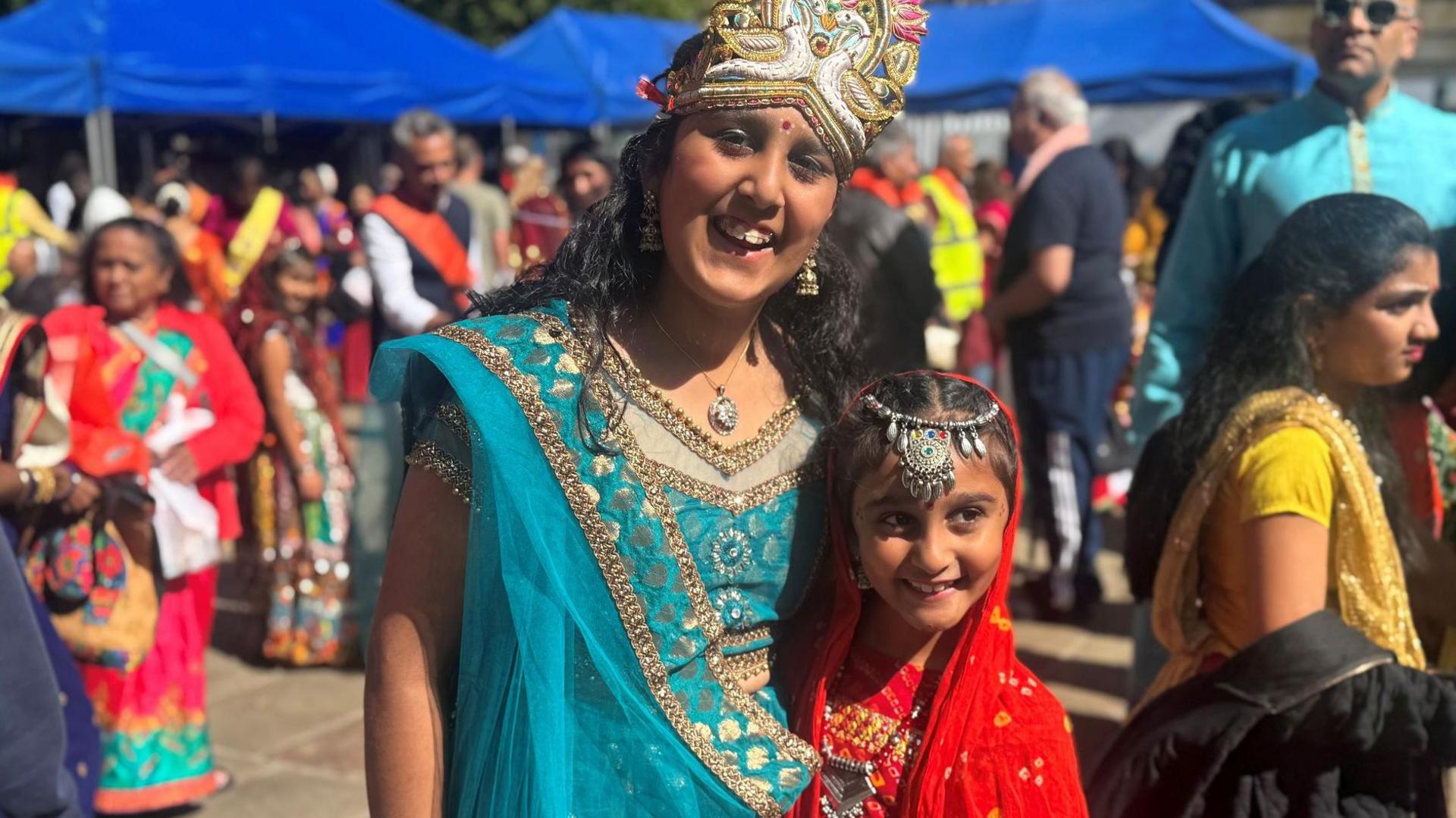 Group images of two girls smiling as they clasp arms at the vent. The older girl wears a sparly multicoloured headdress and red bindi with a sparkly blue sari. The yonger girl wears a red and gold veil and top with silver tiara. Women and mean in colourful Indian outfits can be seen in the background under blue marquees.
