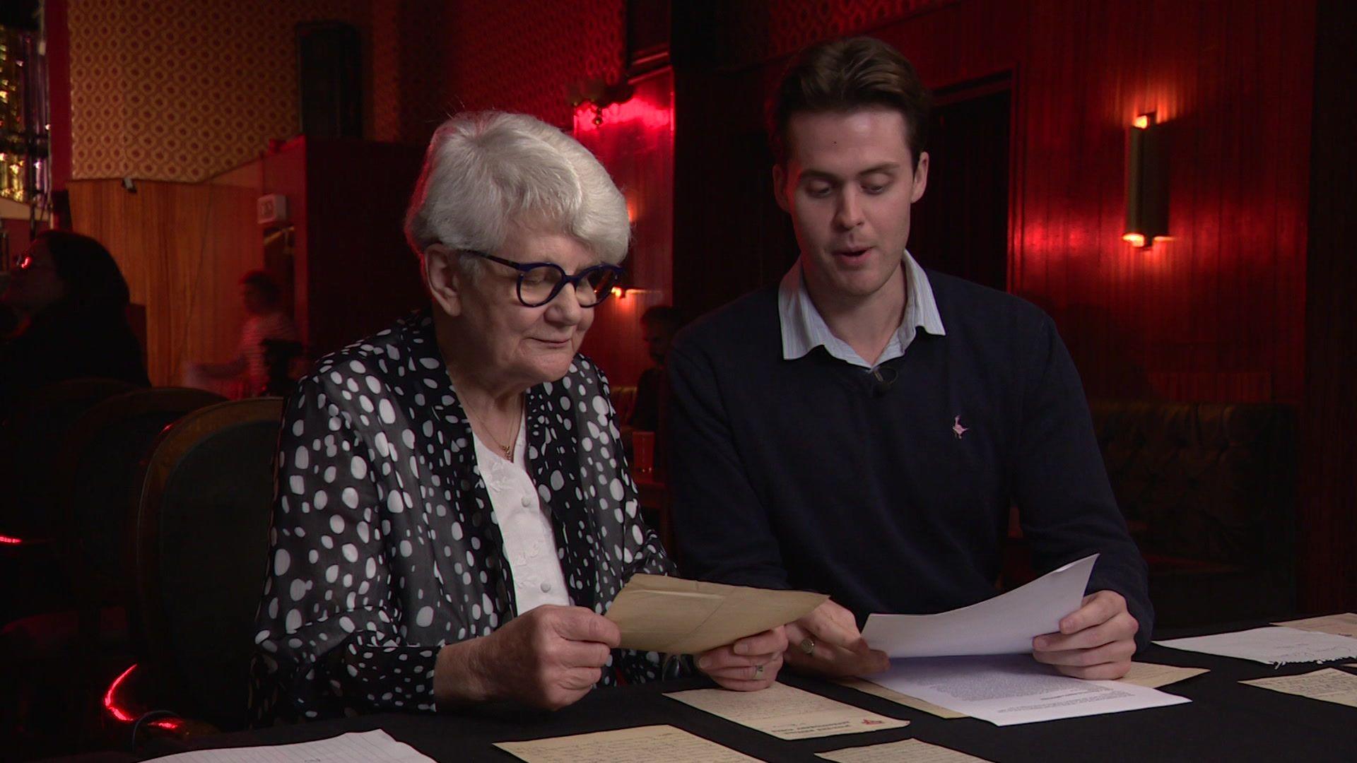 A woman with white short hair, black polka dot blouse and white top underneath, sat at a desk next to her grandson who has a navy jumper, shirt underneath and they're both looking through letters placed piles on the table in front of them.