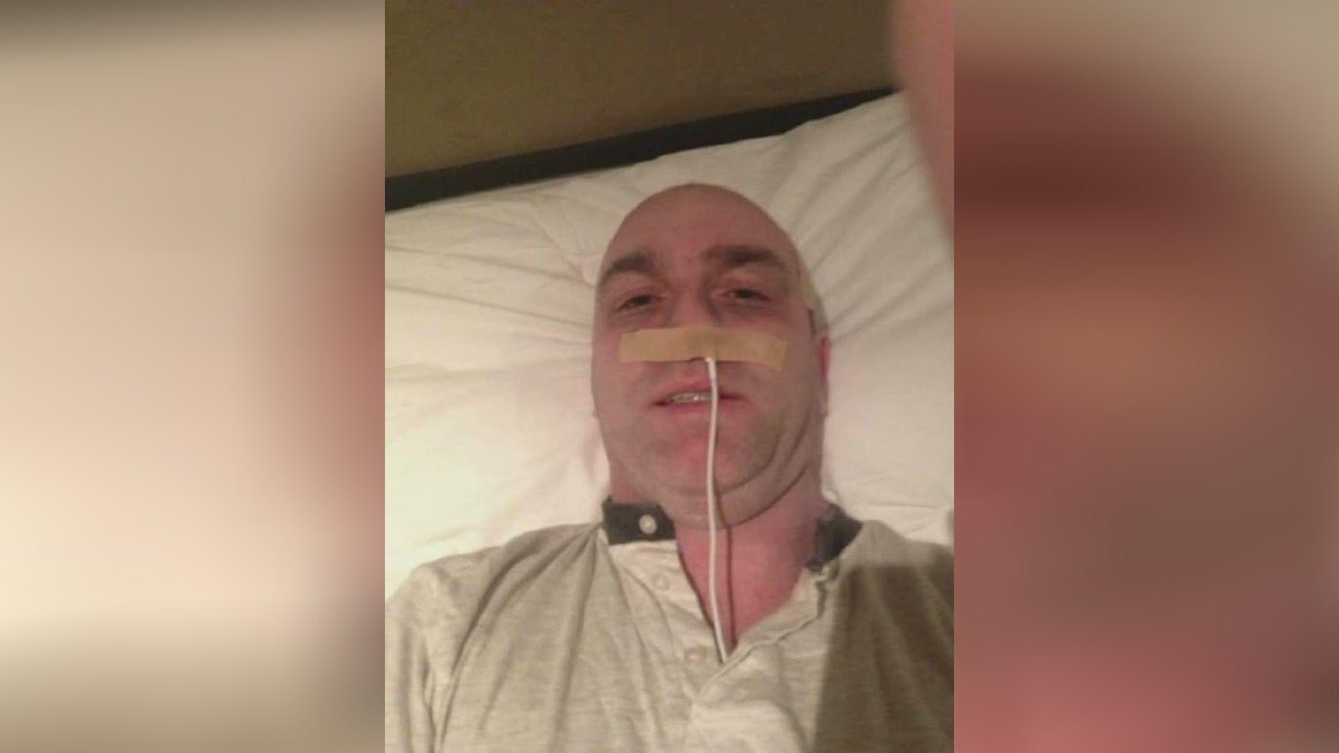 The selfie shows a man lying down on a bed with a white t-shirt on, he has a cord (that is a phone charger lead) up his nose and a piece of light tape over his nose. There is also tape at the sides of his head.