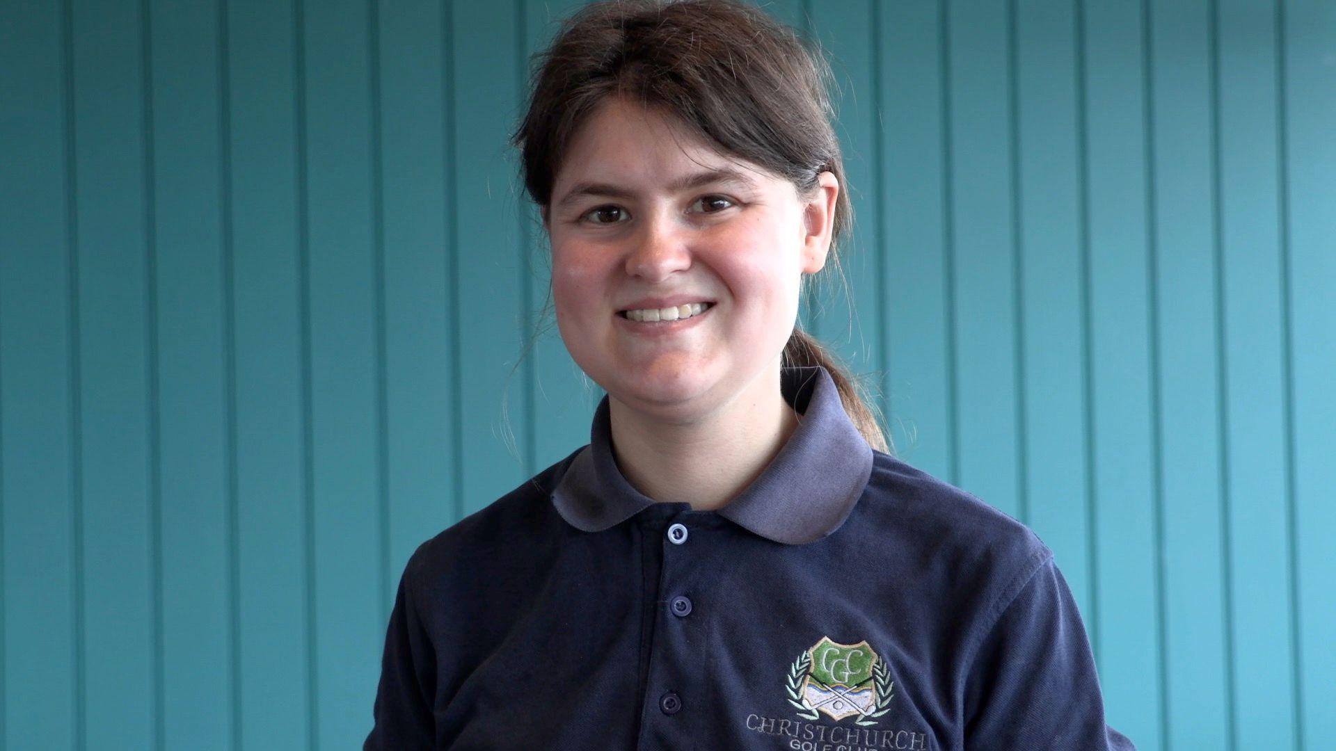 Jade, a white young woman with pale brown hair tied back in a ponytail and wearing a navy blue polo shirt, smiles at the camera with a blue wooden panel background behind her