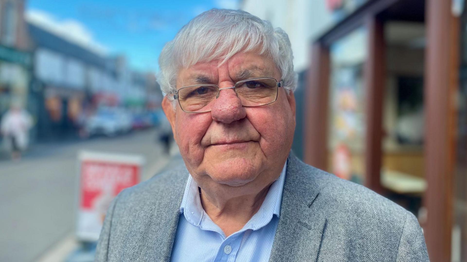 Mr MacKenzie has white hair and glasses. He is standing out in a street wearing a grey tweed jacket over a light blue shirt.