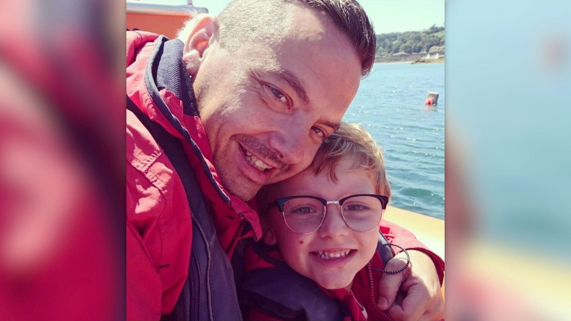 A photo of Dean Lowe, a man with brown hair and stubble wearing a red jacket, hugging Charlie Lowe, a young boy wearing glasses and a red jacket. The sea is in the background behind them.