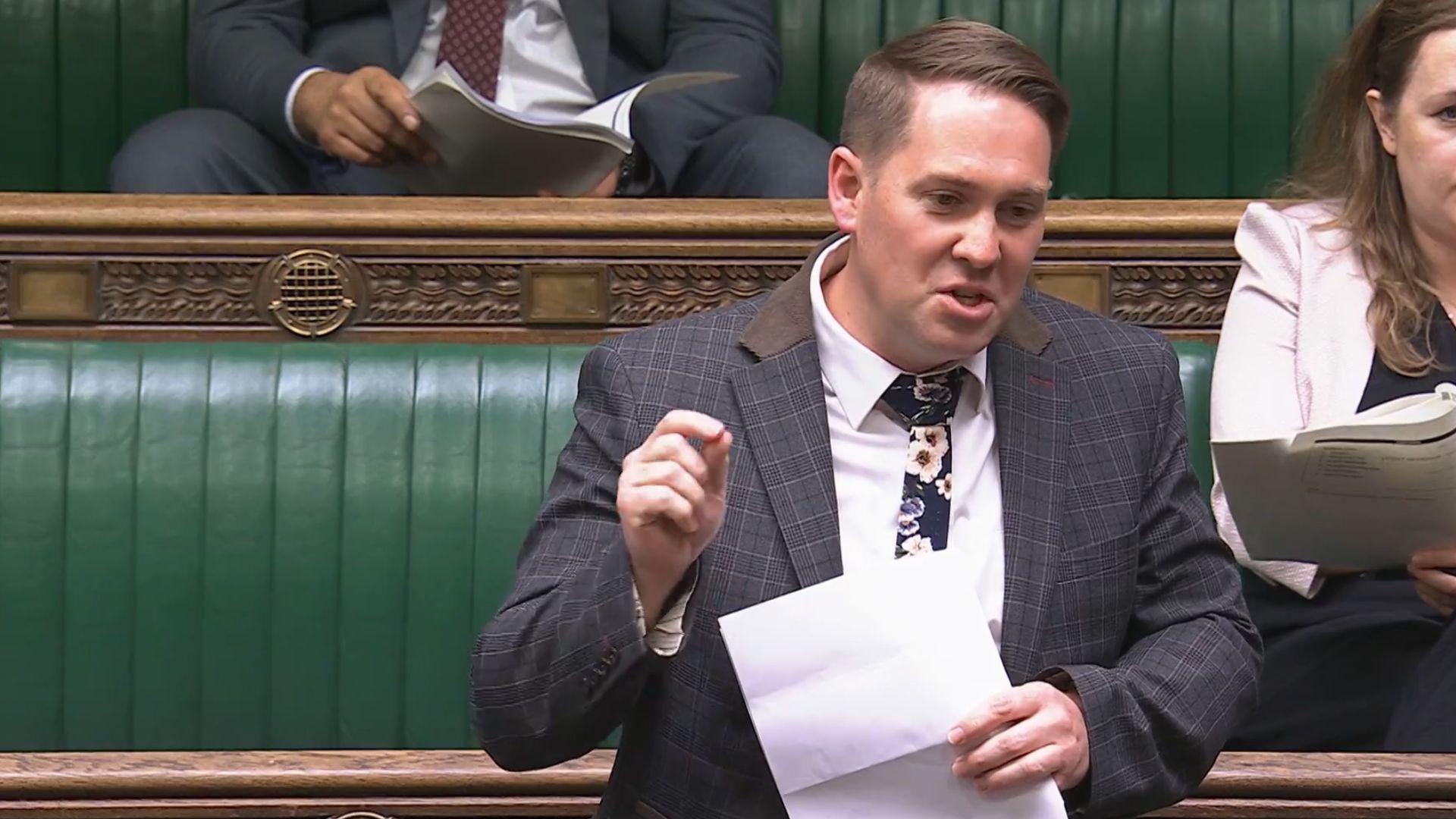 MP Sam Rushworth speaking in the House of Commons. He has dark hair, brushed to his right, and is wearing a dark blazer, white shirt and dark tie with flowers on. Behind him are parliament's green benches where fellow MPs are sitting.