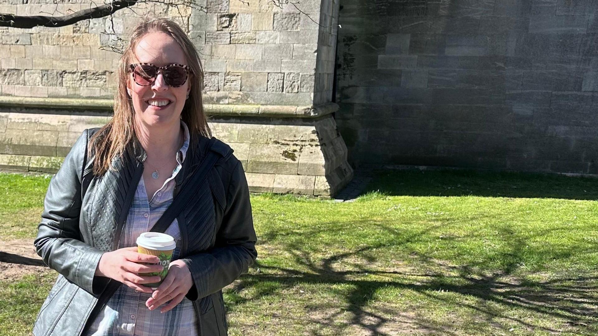 Carla Linford, a woman with long light brown hair, is standing in front of a large stone building. She is smiling for the camera while cupping a takeaway coffee cup.