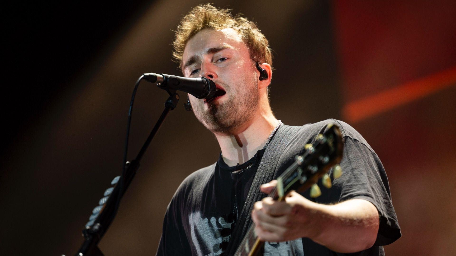 Sam Fender singing and playing the guitar on stage. He's wearing a grey t-shirt with a graphic on the front and is stood in front of a microphone.