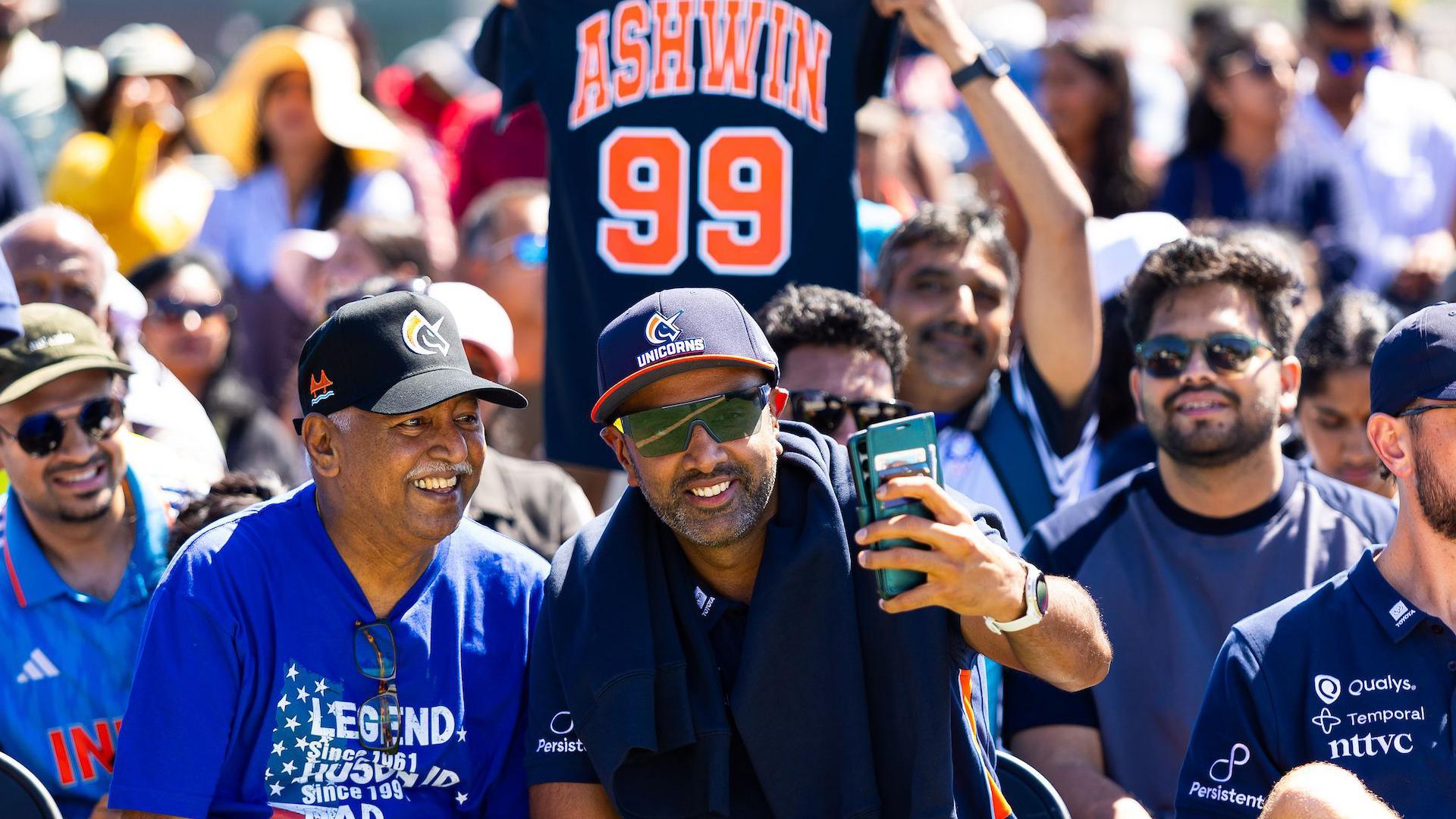 Ravichandran Ashwin is sat with a group of San Francisco Unicorns fans holding a phone in his left hand taking a selfie with them. In the background you see a fan holding up a navy blue shirt with the words Ashwin and number 99 on the back. 