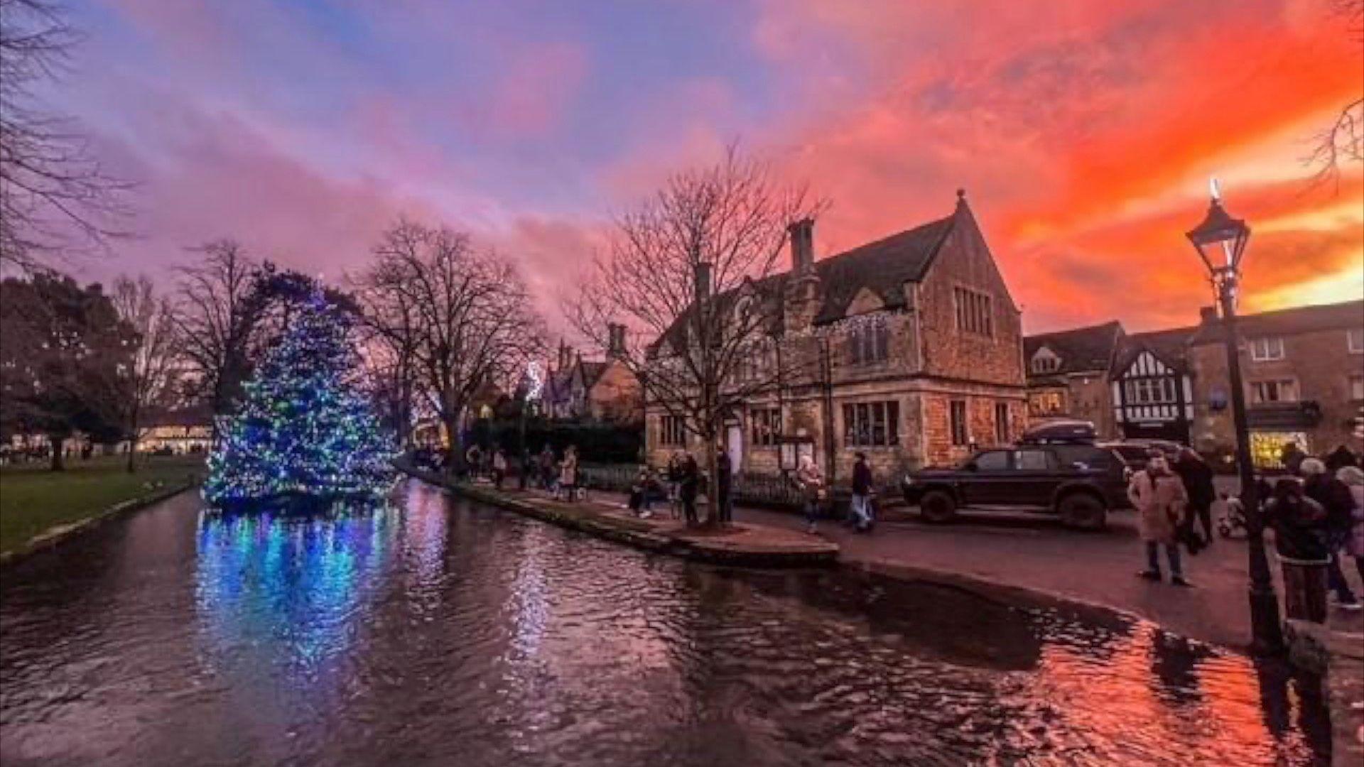 A large Christmas tree on the River Windrush with a pink sky in the background and Cotswold brick buildings. 