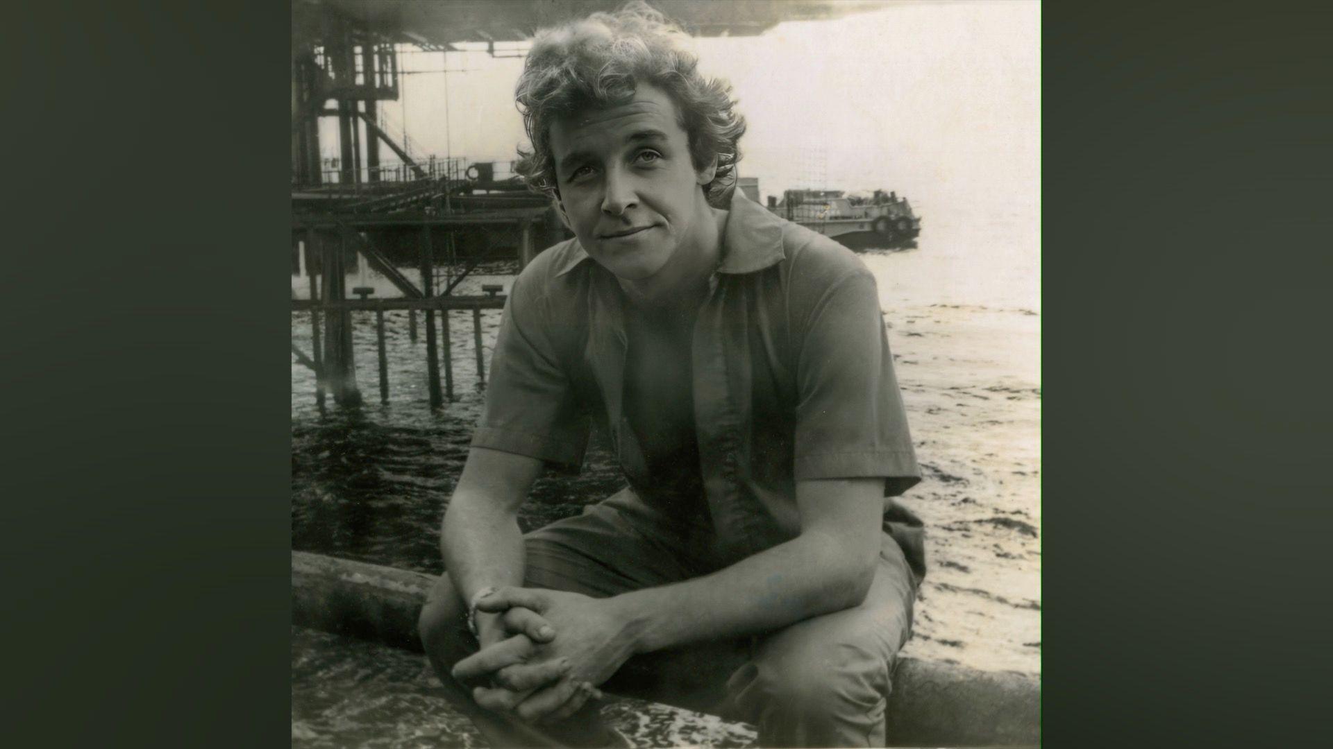 Justin looks at the camera in a black and white photos. His shirt is unbuttoned and his hands are crossed. He's sitting by  the seaside with a pier in the background.