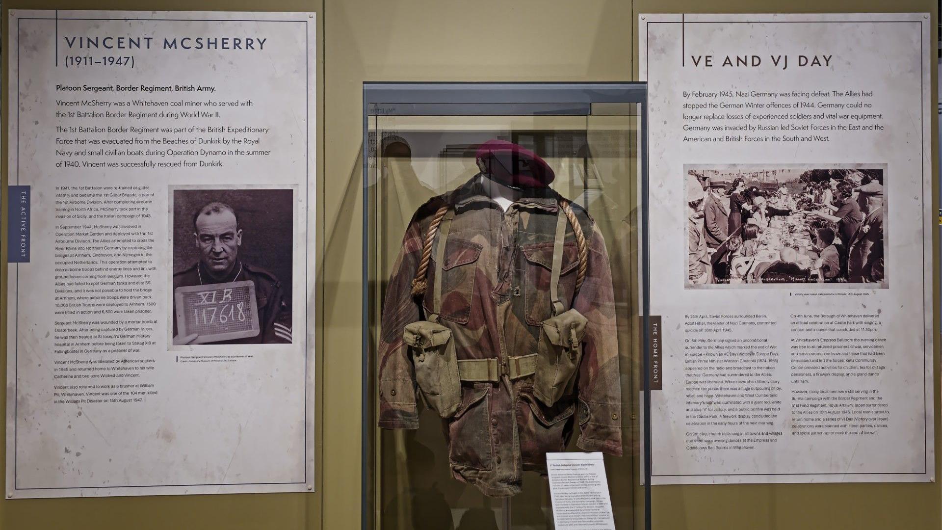 A section of the display. A camouflaged uniform from World War Two is displayed on a mannequin in a glass display. Two signs have been hung on either side of the case. One provides information about a man called Vincent McSherry, the other about VE an VJ Day.