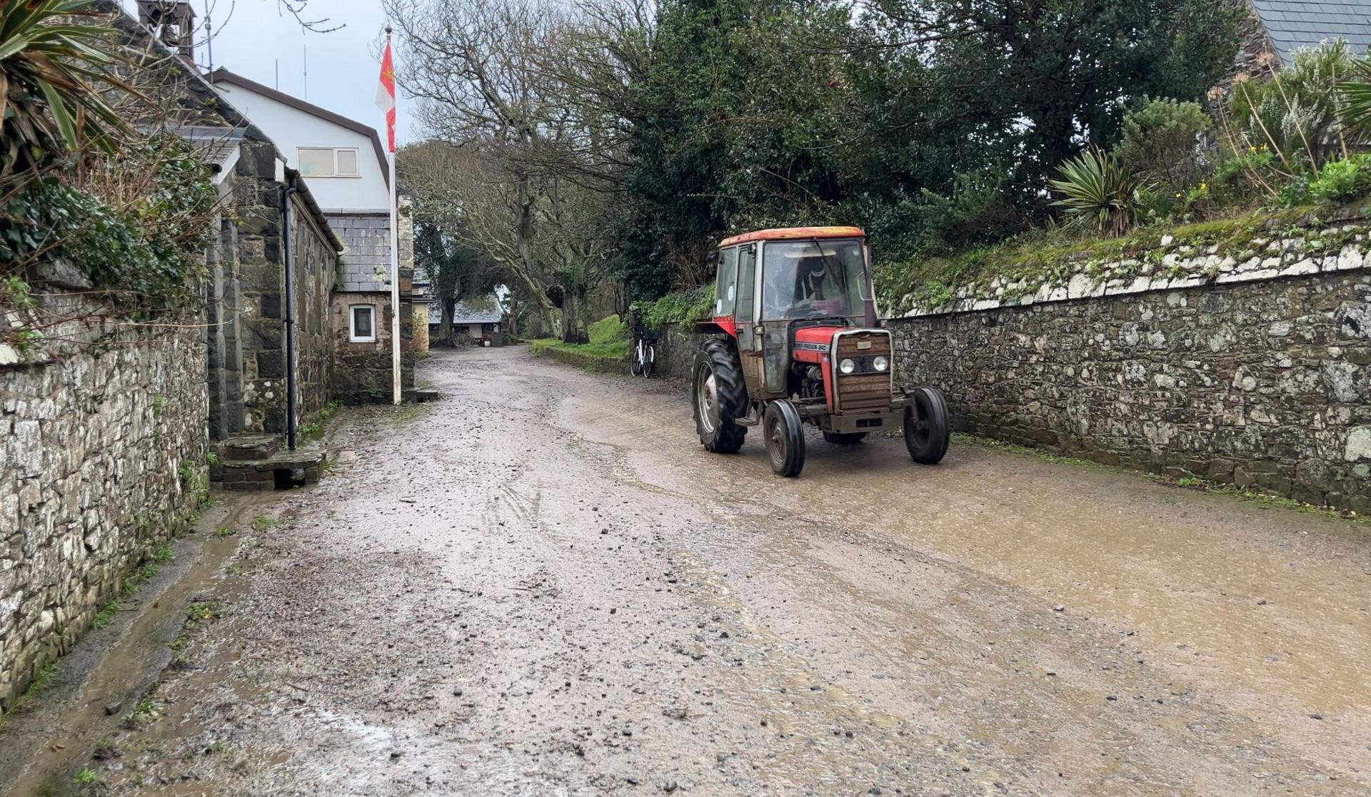 A very muddy road in Sark, with an old-fashioned red tractor on the right-hand side. There are stone walls on either side of the road with shrubbery and trees behind them. There is a building with a flag in front of it to the left.