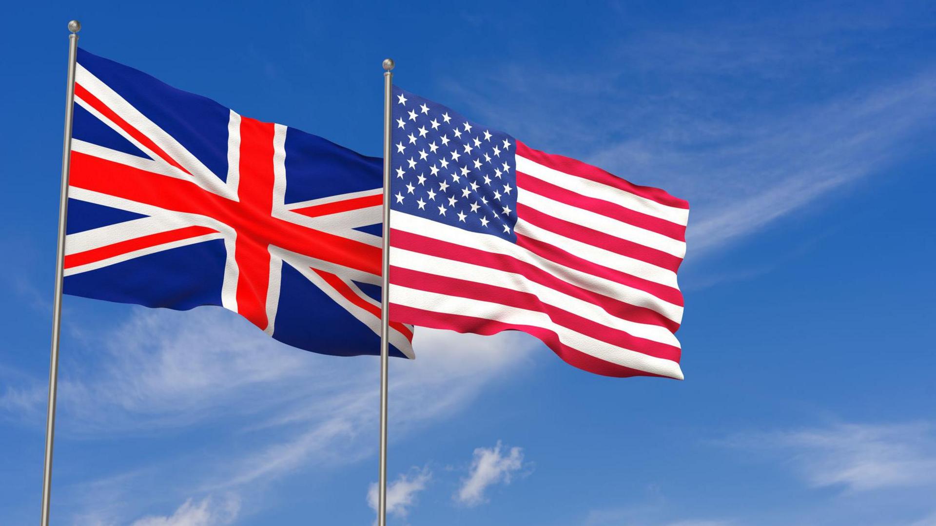 The US and the UK flags fly on flagpoles, against the backdrop of a blue sky