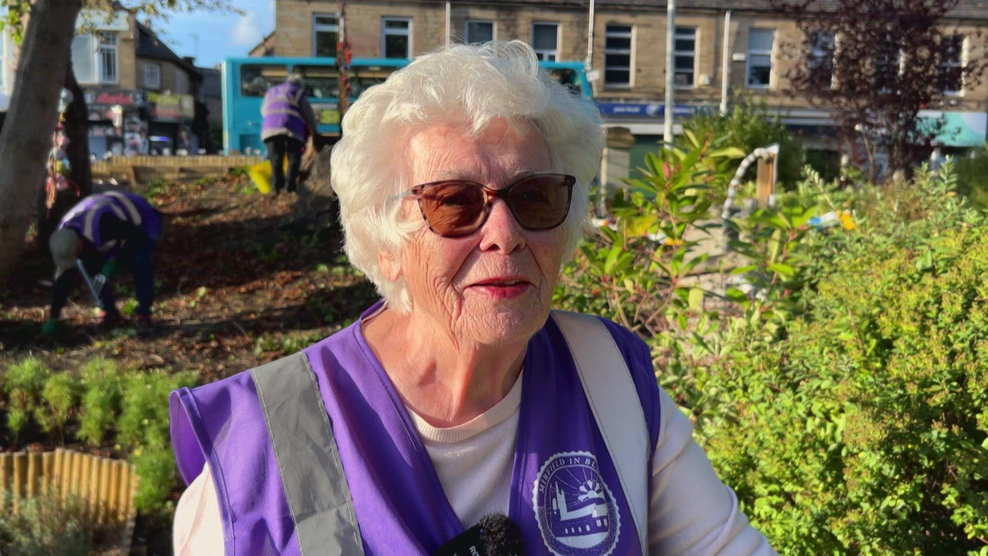 A woman with white hair, wearing sunglasses. She is wearing a purple Mirfield in Bloom overall and is standing in front of some bushes and shrubs with other volunteers and shops in the background. 
