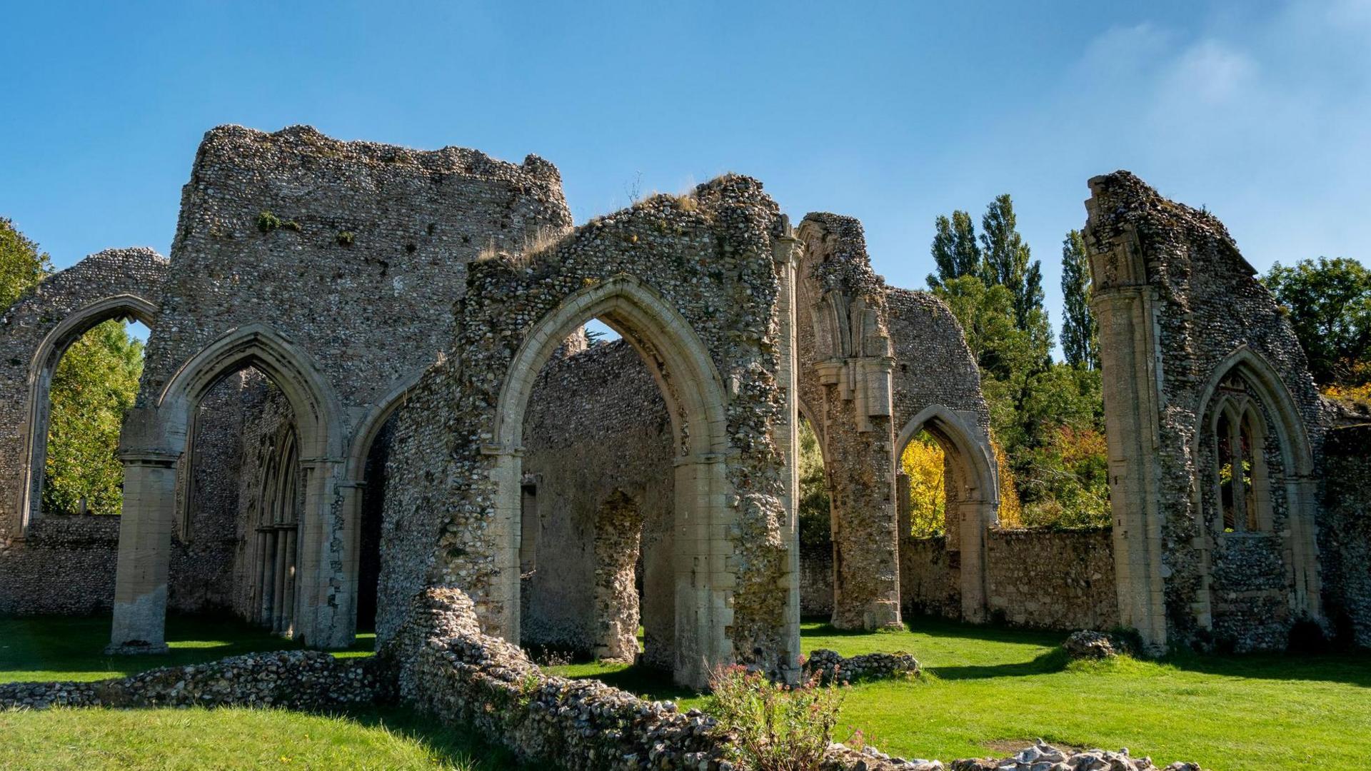 The ruins of Creake Abbey, showing cream stone arches and some broken down flint-built walls under a blue sky. The site is well-maintained with short cropped grass.