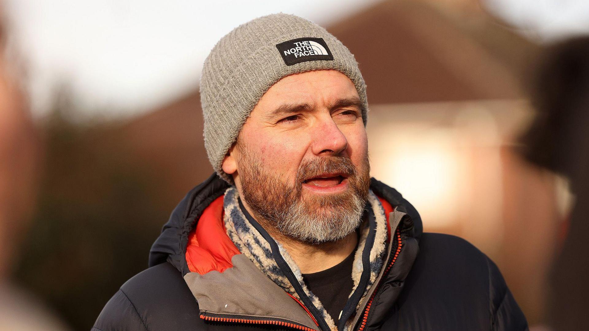 Steve North, wearing a beige beanie hat, leopard-print scarf and blue coat, speaks outside the protest