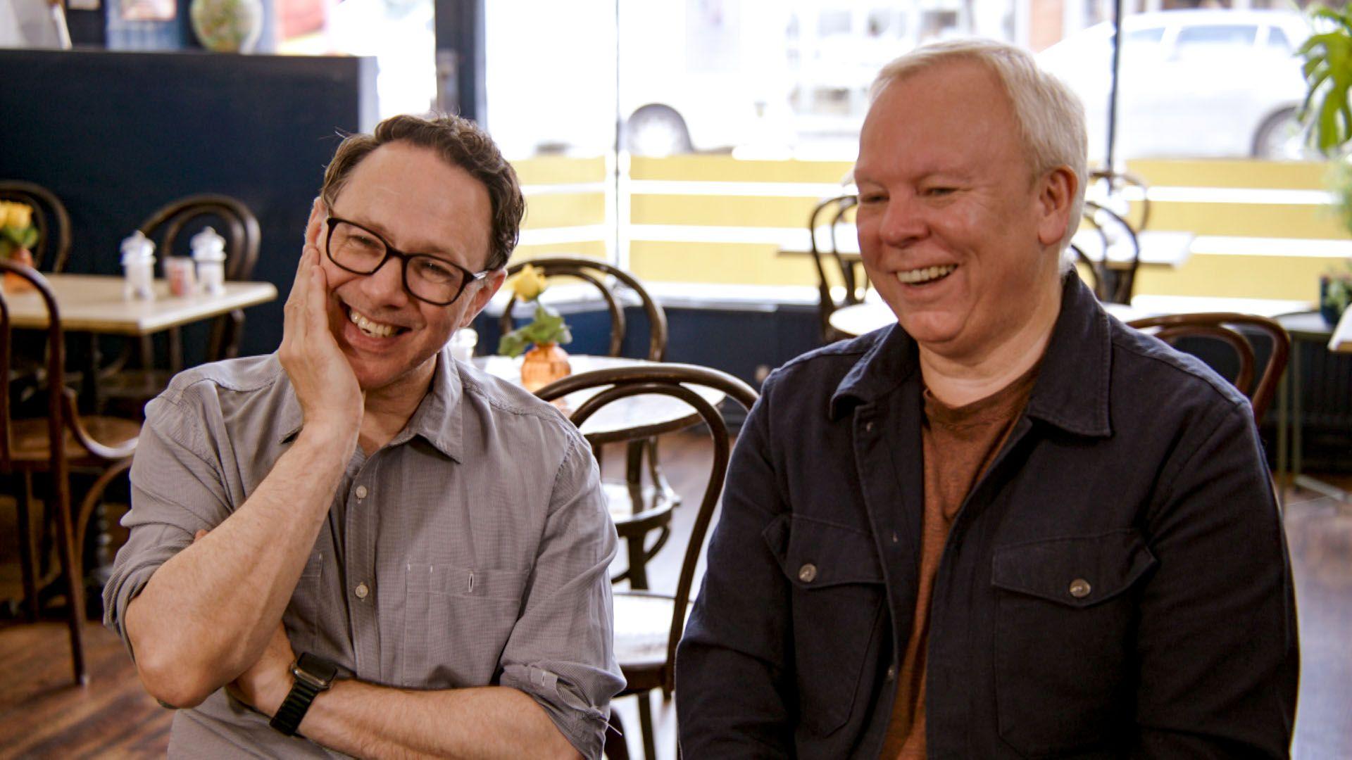 The image shows two men sat indoors in a café area. They are sitting side by side on wooden chairs with curved backs. The man on the left wears a light grey button-up shirt with rolled up sleeves and has one hand resting against the side of their face. The man on the right is wearing a dark jacket over a brown shirt. In the background, there are several empty tables with wooden chairs, small vases of flowers, and salt and pepper pots. Large windows behind them let in light.