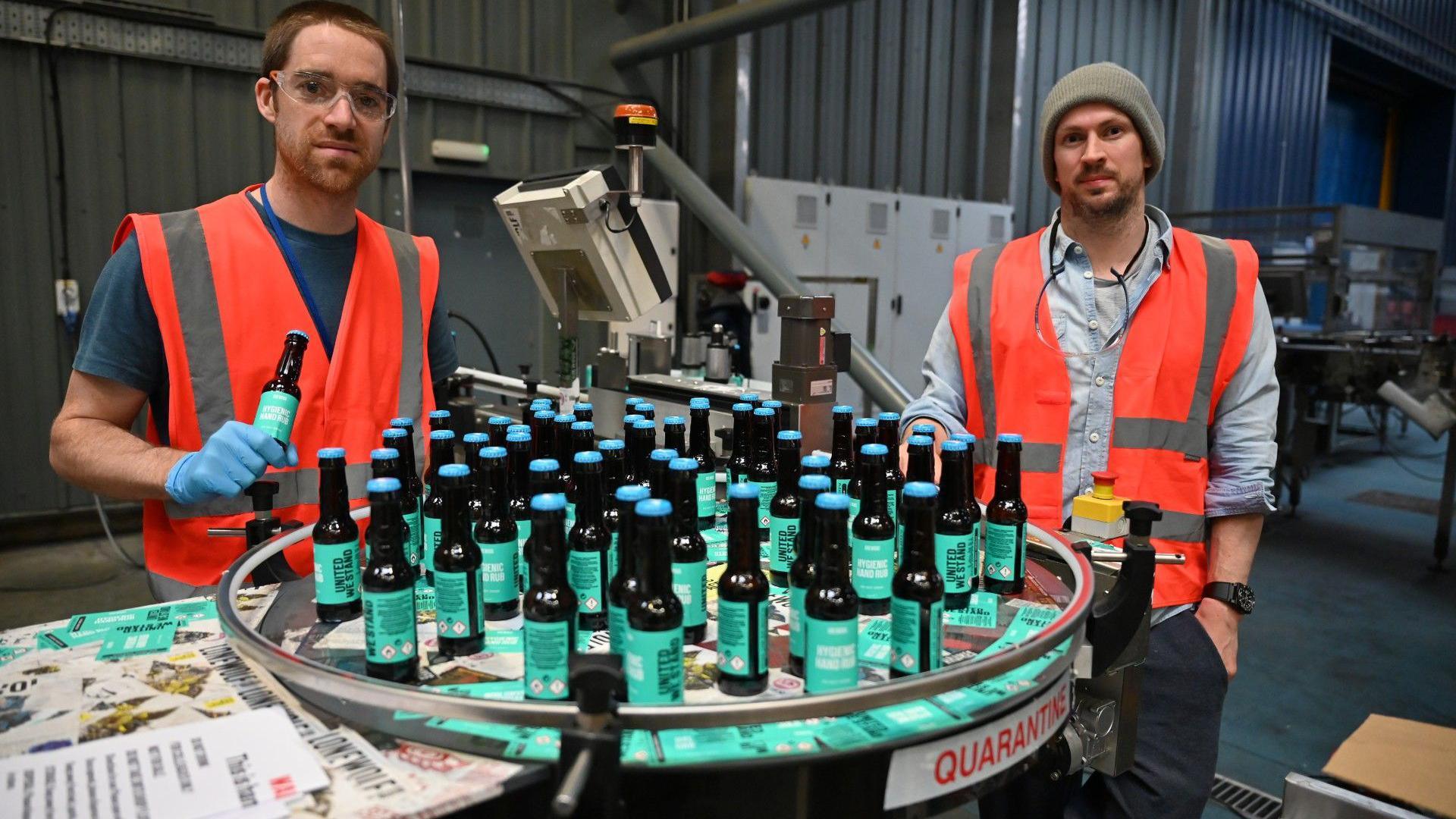 Martin Dickie - who has short brown hair and heavy stubble - and an unshaven James Watt, who is wearing a grey beanie hat, (right) pictured packing bottles of BrewDog hand sanitizer during the Covid pandemic in April 2020. The black bottles have green labels and light blue tops. Both men are wearing orange hi-viz vests. Dickie is also wearing clear safety glasses.