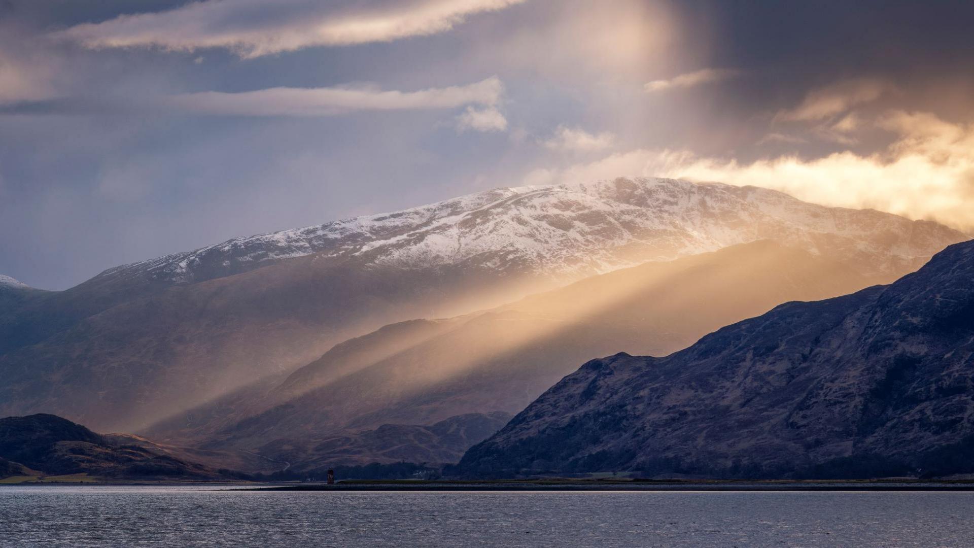 Shafts of sunlight on hills dusted with snow and the loch in the foreground.