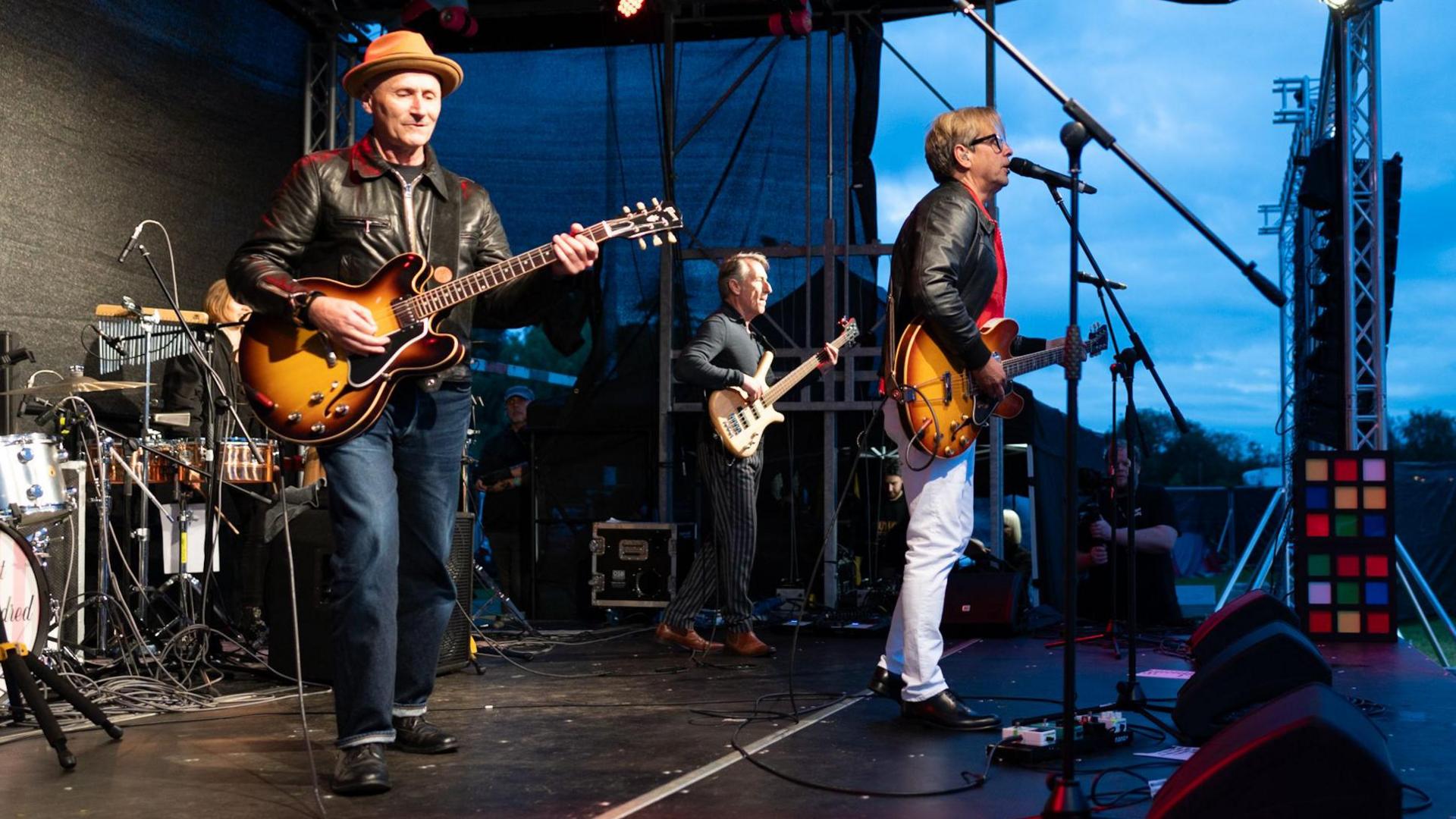 Three members of the band Haircut 100, each holding a guitar and wearing a black leather jacket, stand slightly apart on a stage, as one sings into a microphone