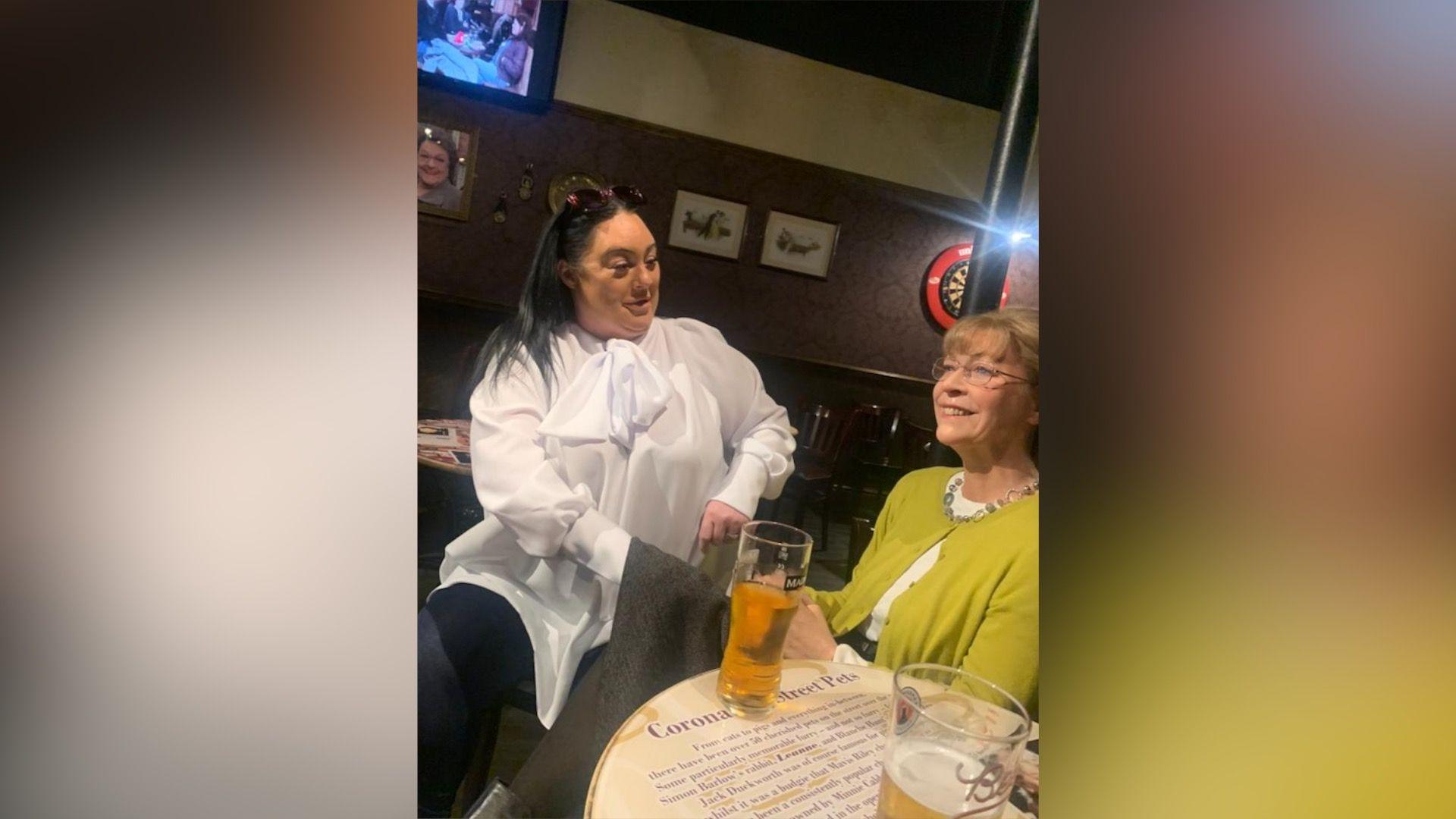 Michelle Robinson sits with a waxwork of Deirdre Barlow in a replica of the Rovers Return Inn. She wears a white shirt and black leggings