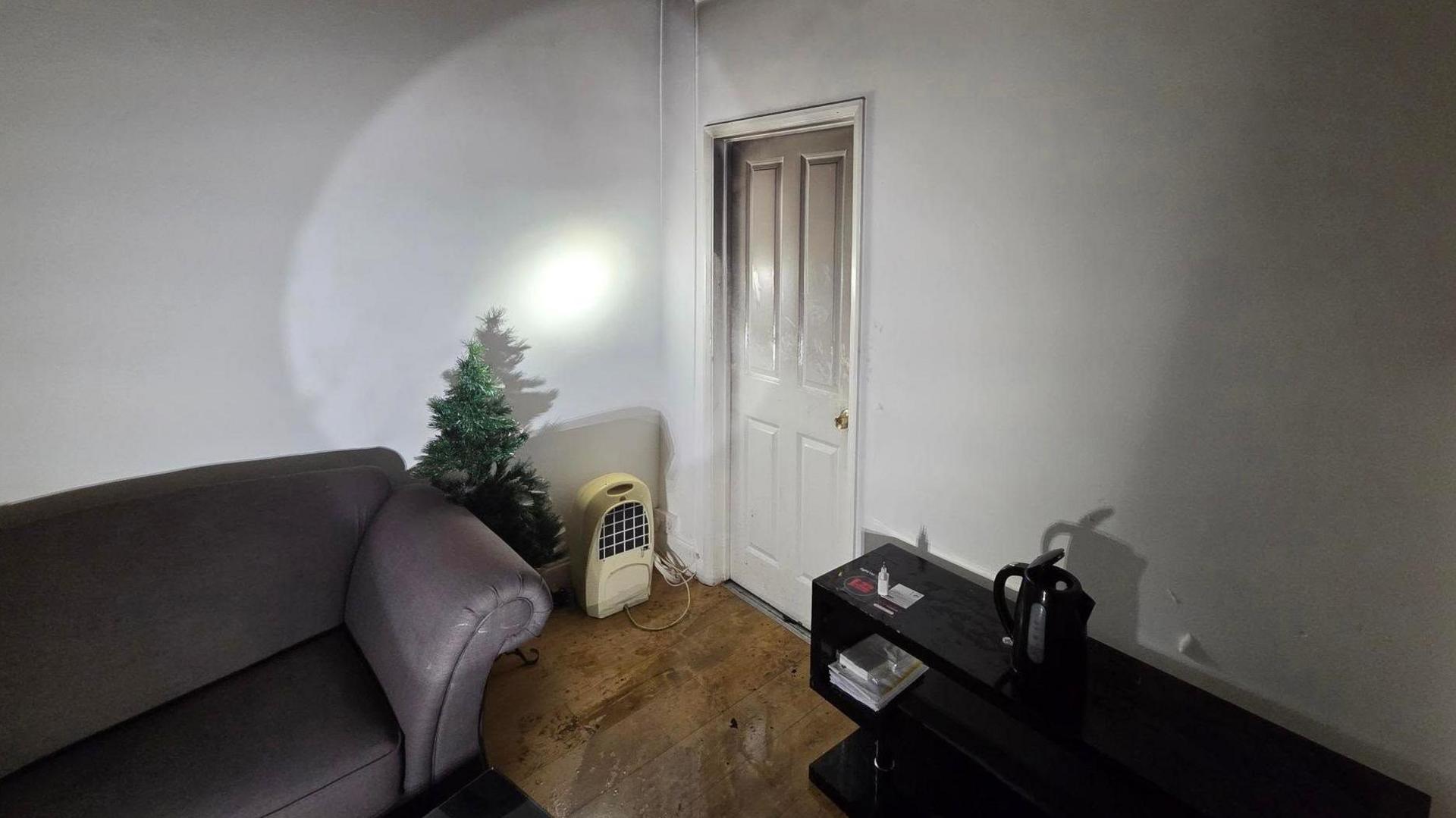Sitting room with door closed on right hand side, a small Christmas tree and grey sofa are on the back wall and a low, black sideboard is along the right wall.