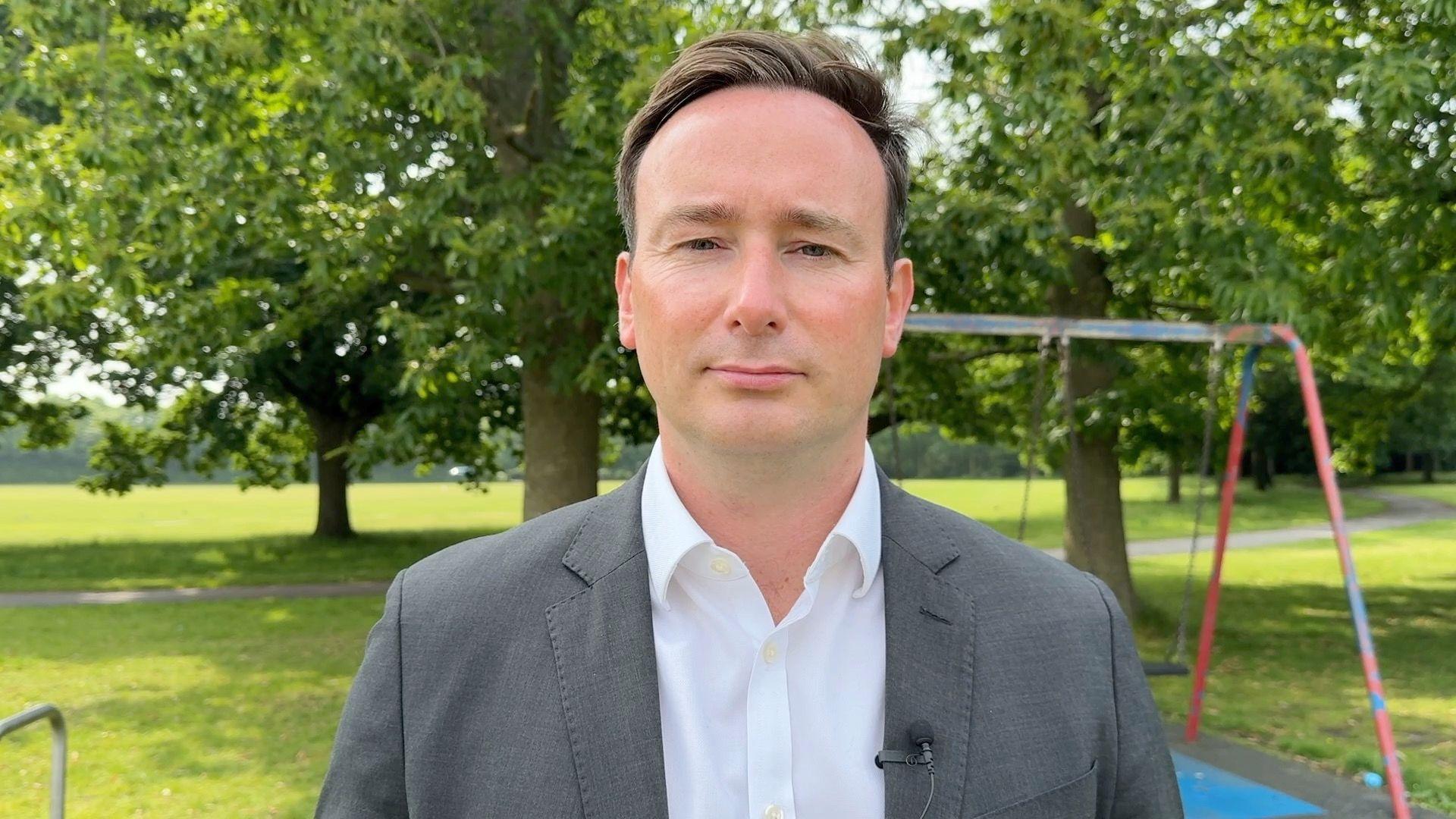 MP for Bournemouth East Tom Hayes. He is stood in a park. Behind him are lots of trees. There is also a red and blue swing behind his right shoulder. He is wearing a white shirt with a grey suit jacket. 