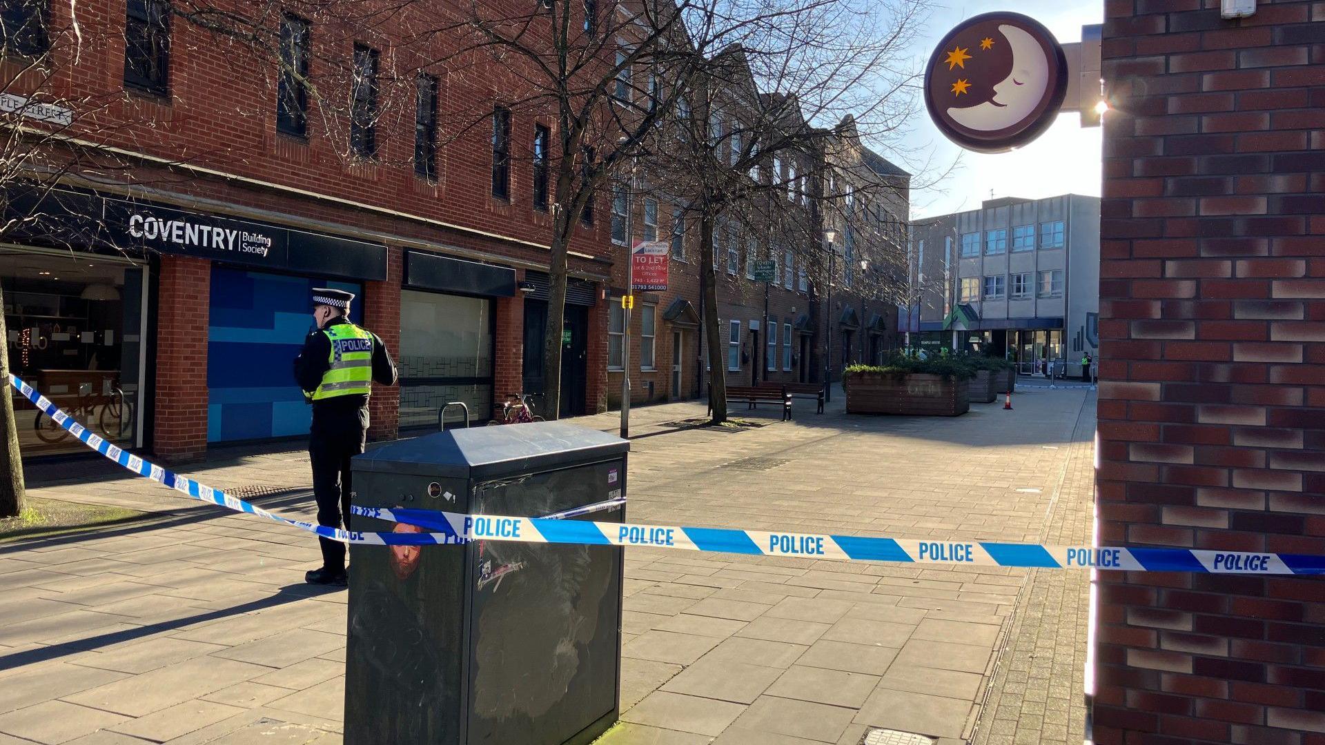 A police officer is standing next to police tape in a town centre.