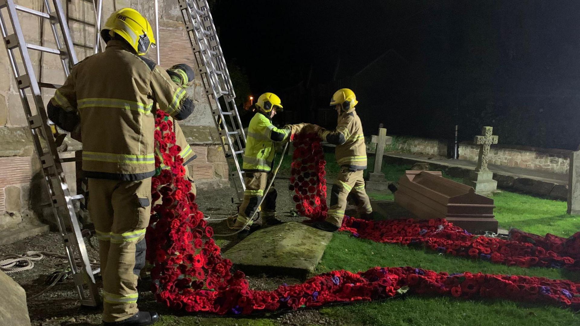 Three firefighters wearing full kit - beige coats and trousers with yellow reflective material, and yellow helmets. They're holding the ends of the poppy cascade and are about to climb two metal ladders propped up against the church bell tower. It's pitch black outside, so it taken late at night, but flood lights have been set up. 